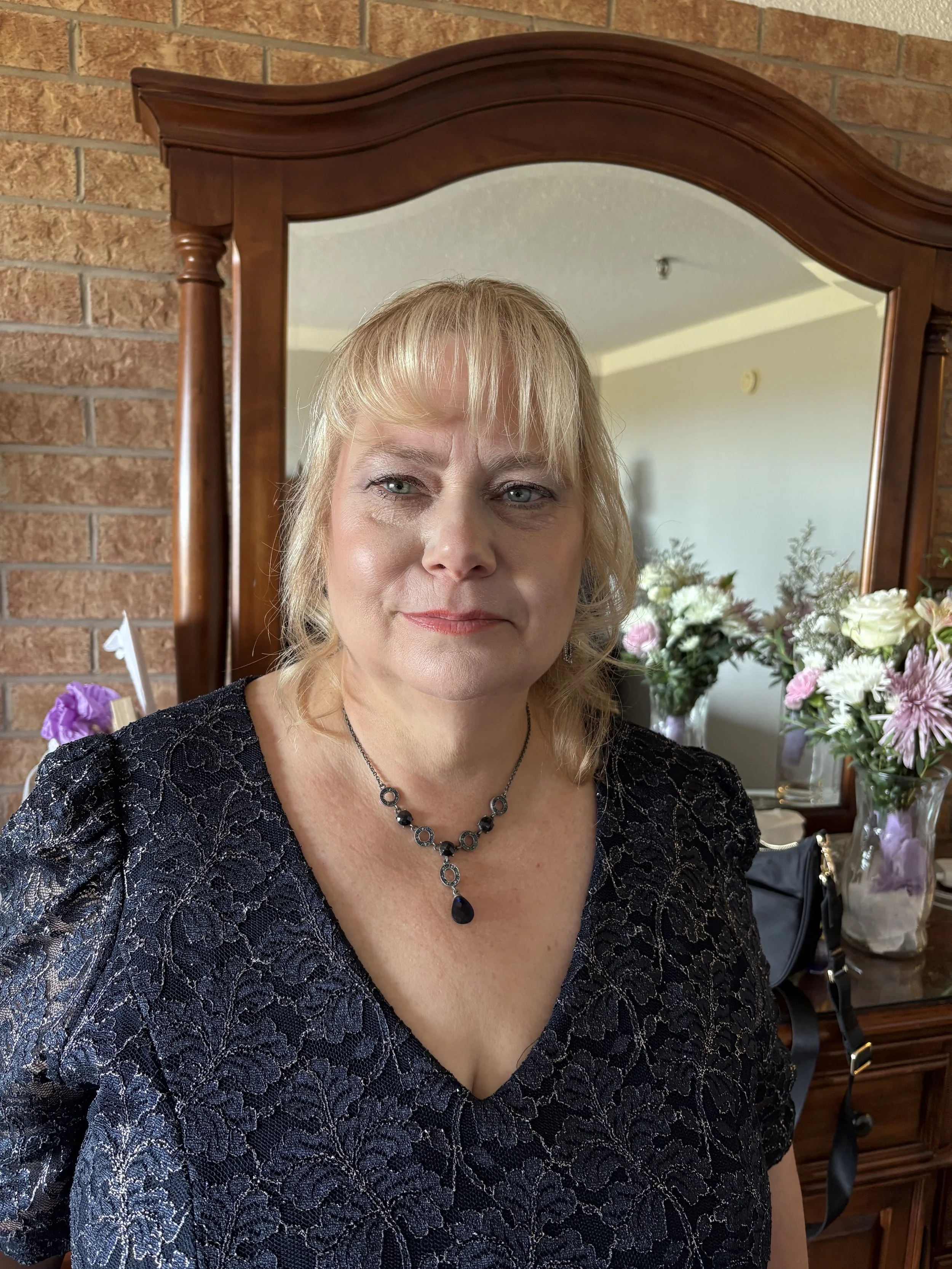 A woman with blonde hair and blue eyes posing in front of a mirror, wearing a black lace dress and a black and silver necklace, with a brick wall and floral arrangements in the background.