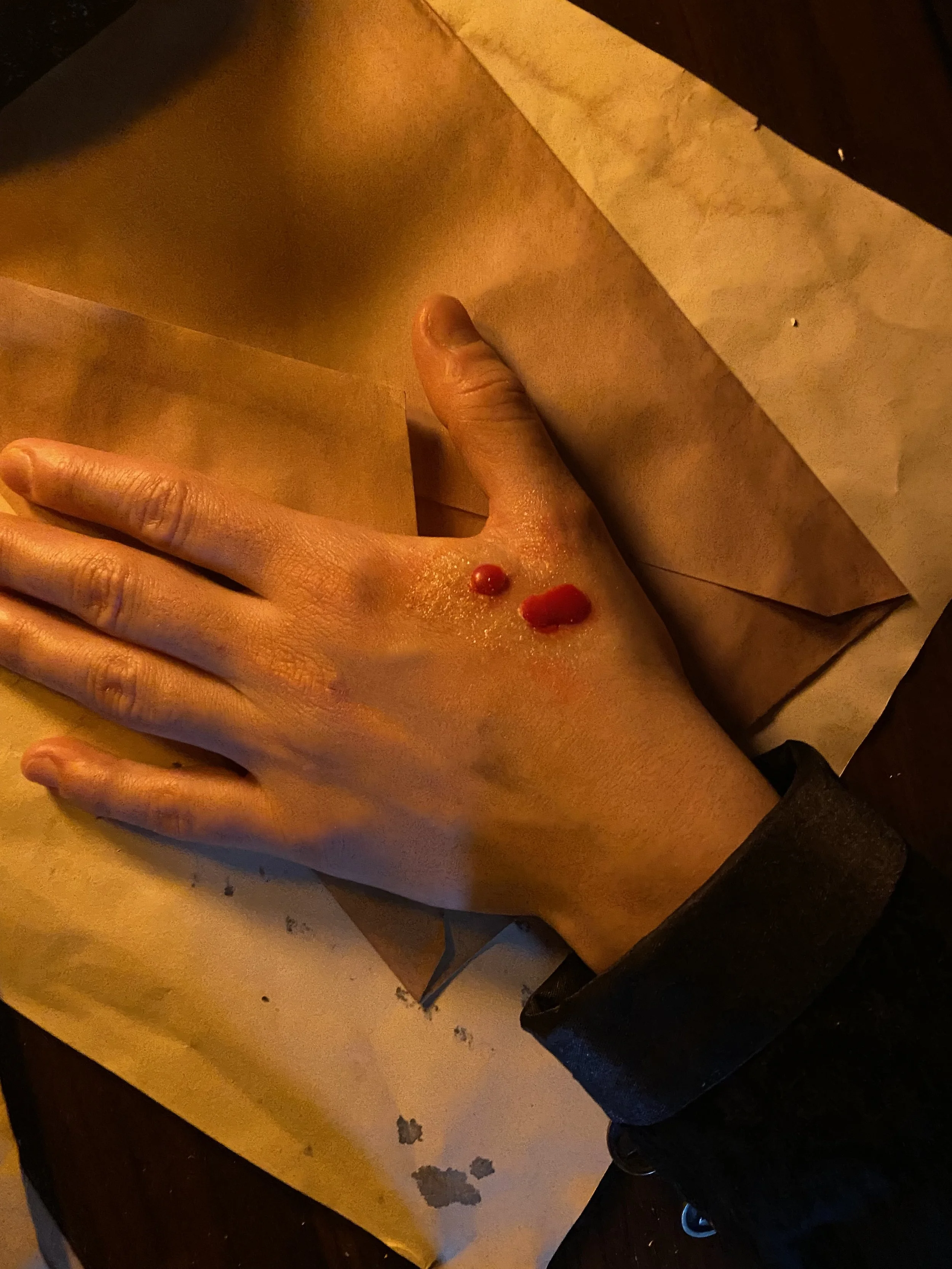 A hand with a small, fresh bleeding cut on the back, resting on brown paper.