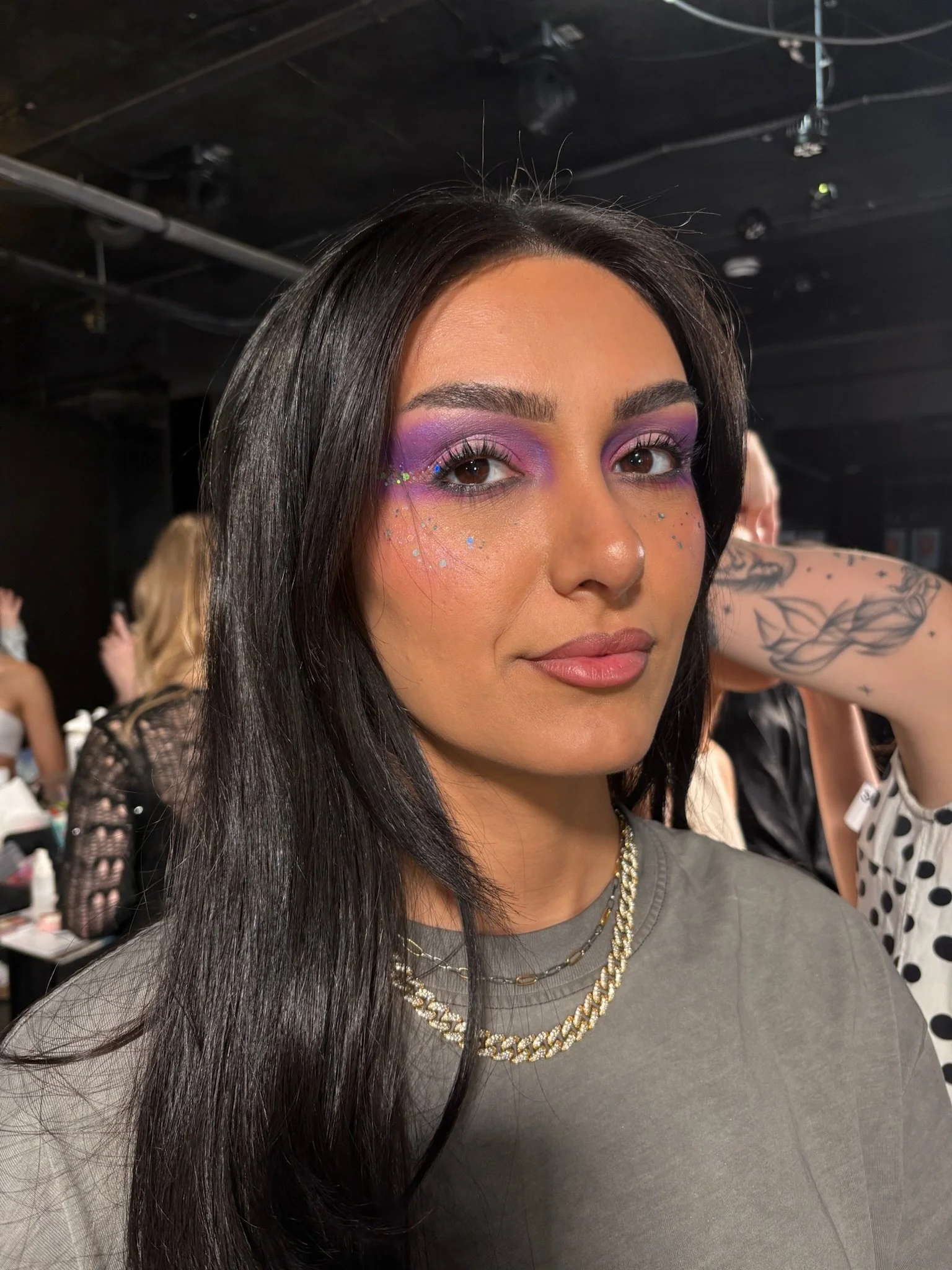 Close-up of a woman with long black hair, purple eye makeup with sparkles, and a neutral expression, in a backstage or studio setting with other people in the background.