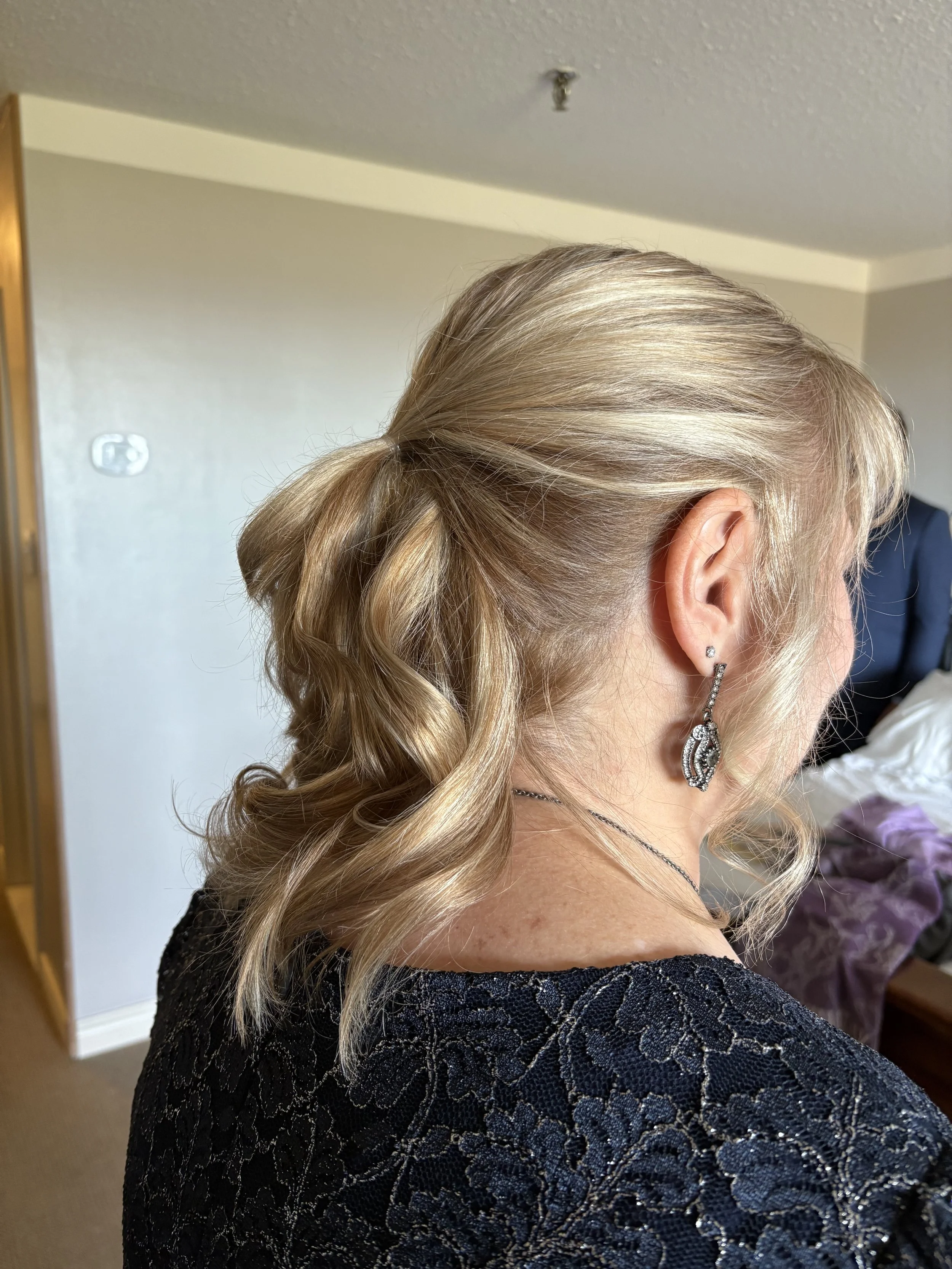 A woman with blonde, curled hair wearing a black lace dress, silver earrings, and a thin necklace, standing indoors with a bed and purple blanket in the background.