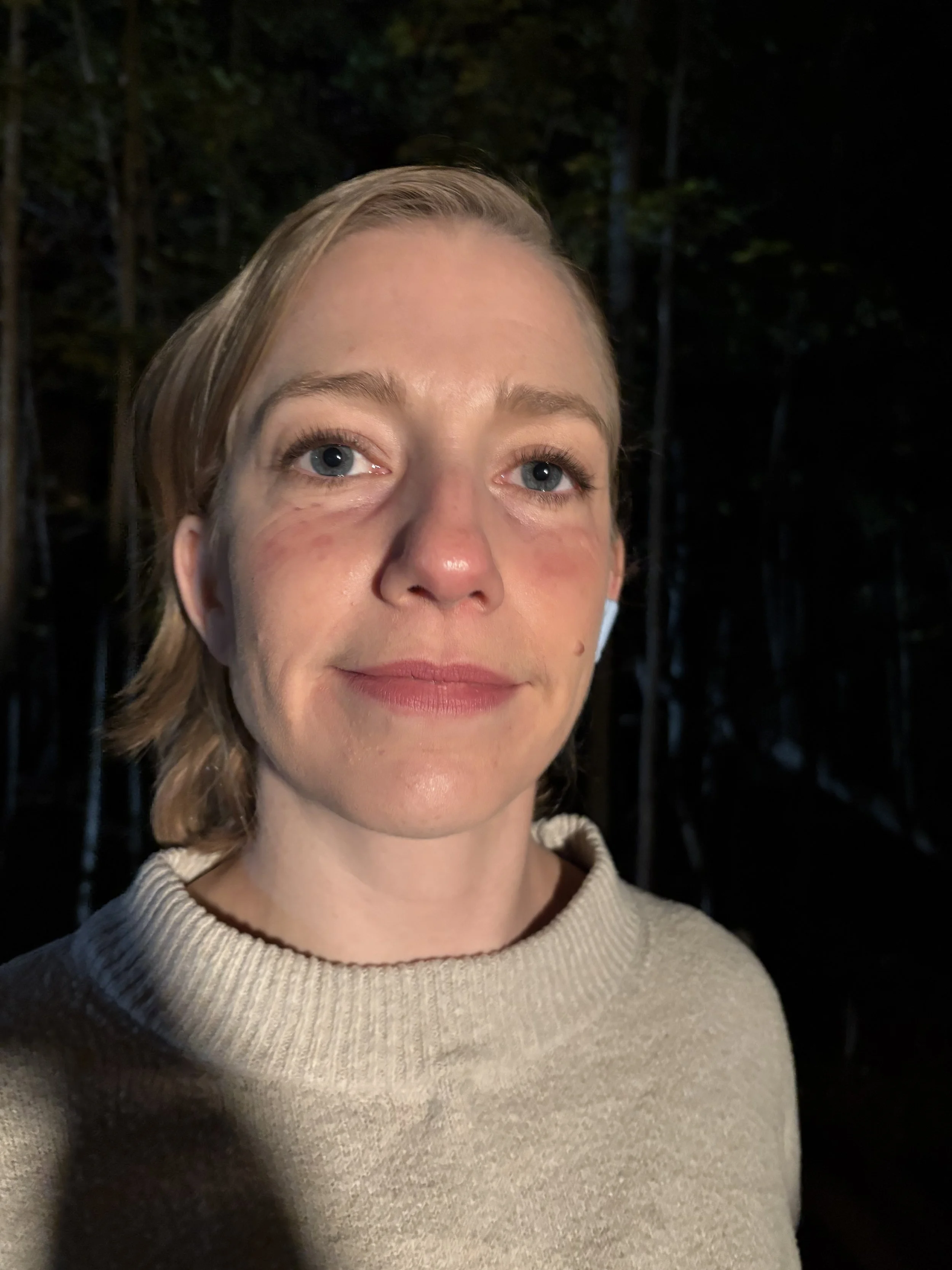 Close-up of a woman with blonde hair and blue eyes, wearing a beige sweater, standing outdoors at night in a wooded area.