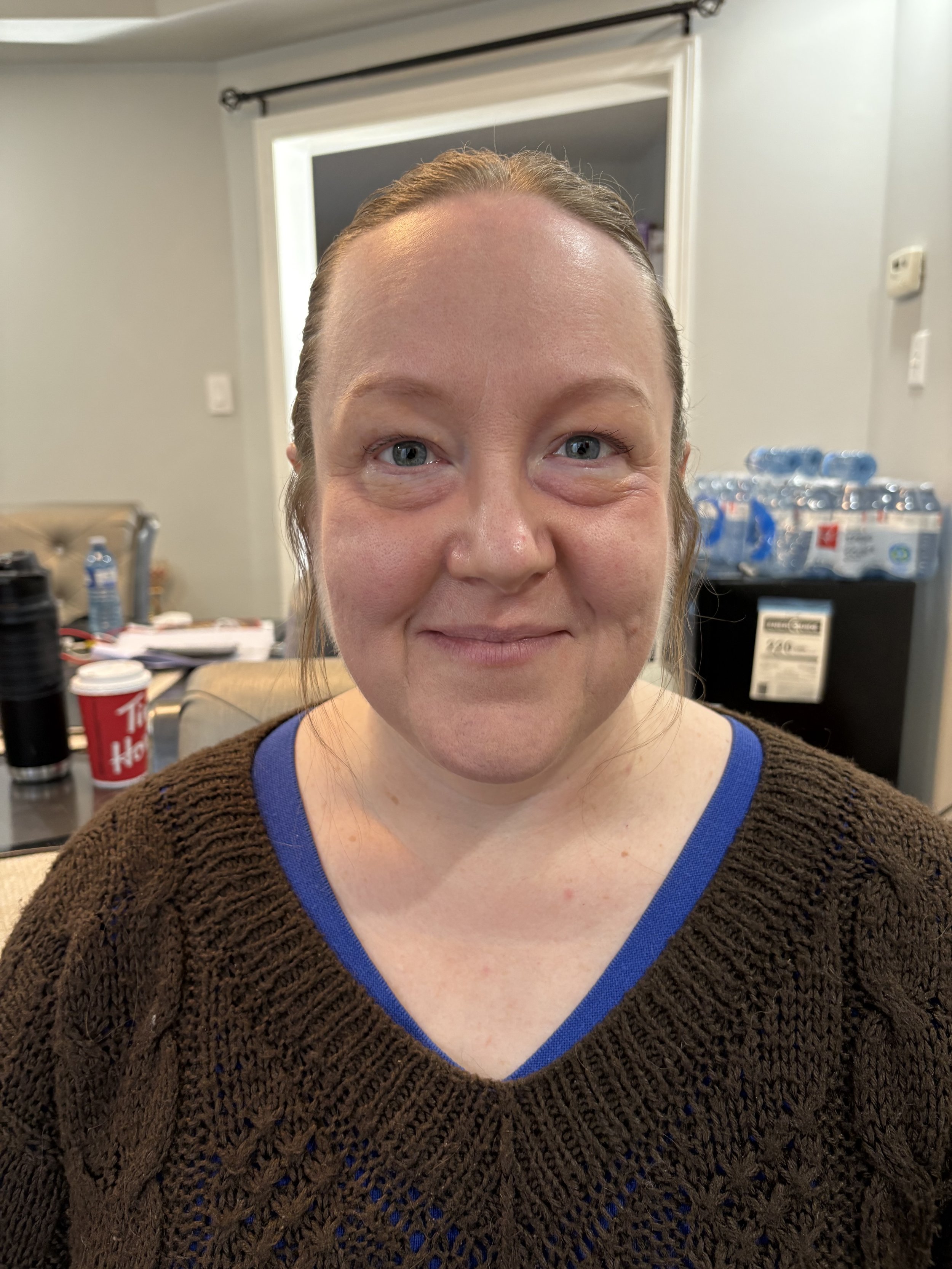 A close-up of a smiling woman with light skin, light blue eyes, and brown hair, wearing a brown sweater with a blue neckline, in an indoor setting with a couch, bottled water, and a Tim Hortons cup visible in the background.