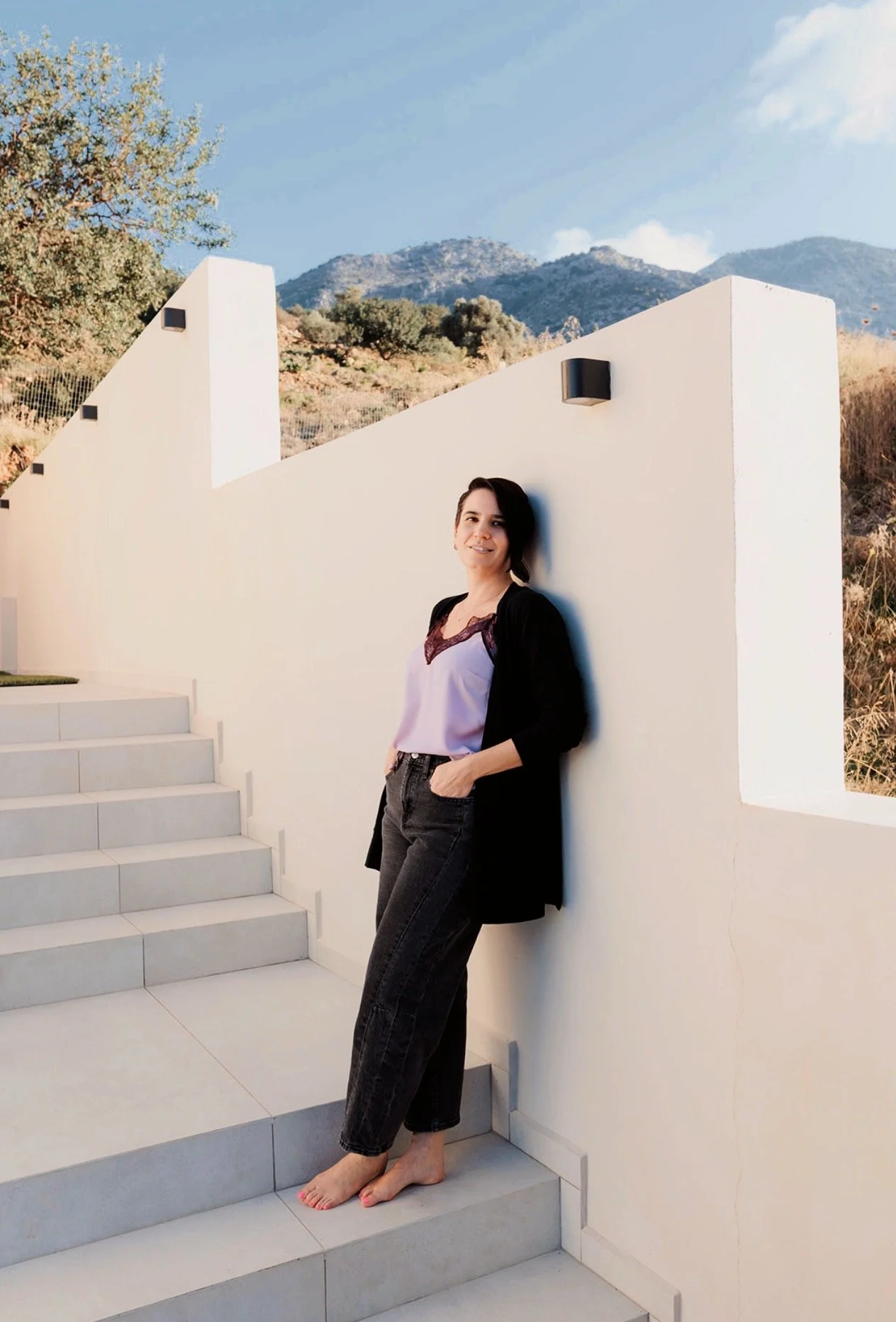 A woman with short black hair, wearing a black jacket, white top, and black jeans, is barefoot, standing on white stairs and leaning against a white wall outdoors. There are mountains and trees in the background, with a clear blue sky.