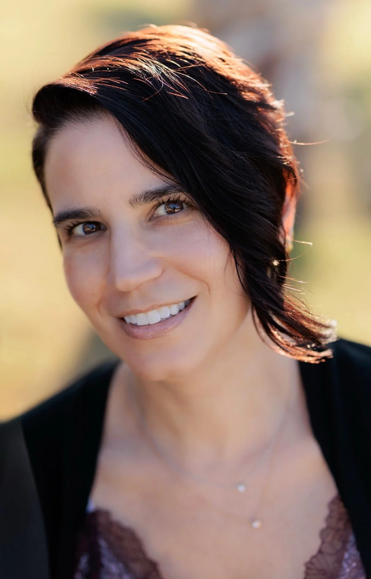 A woman smiling outdoors with short dark hair illuminated by sunlight, wearing a black top, and a delicate necklace.