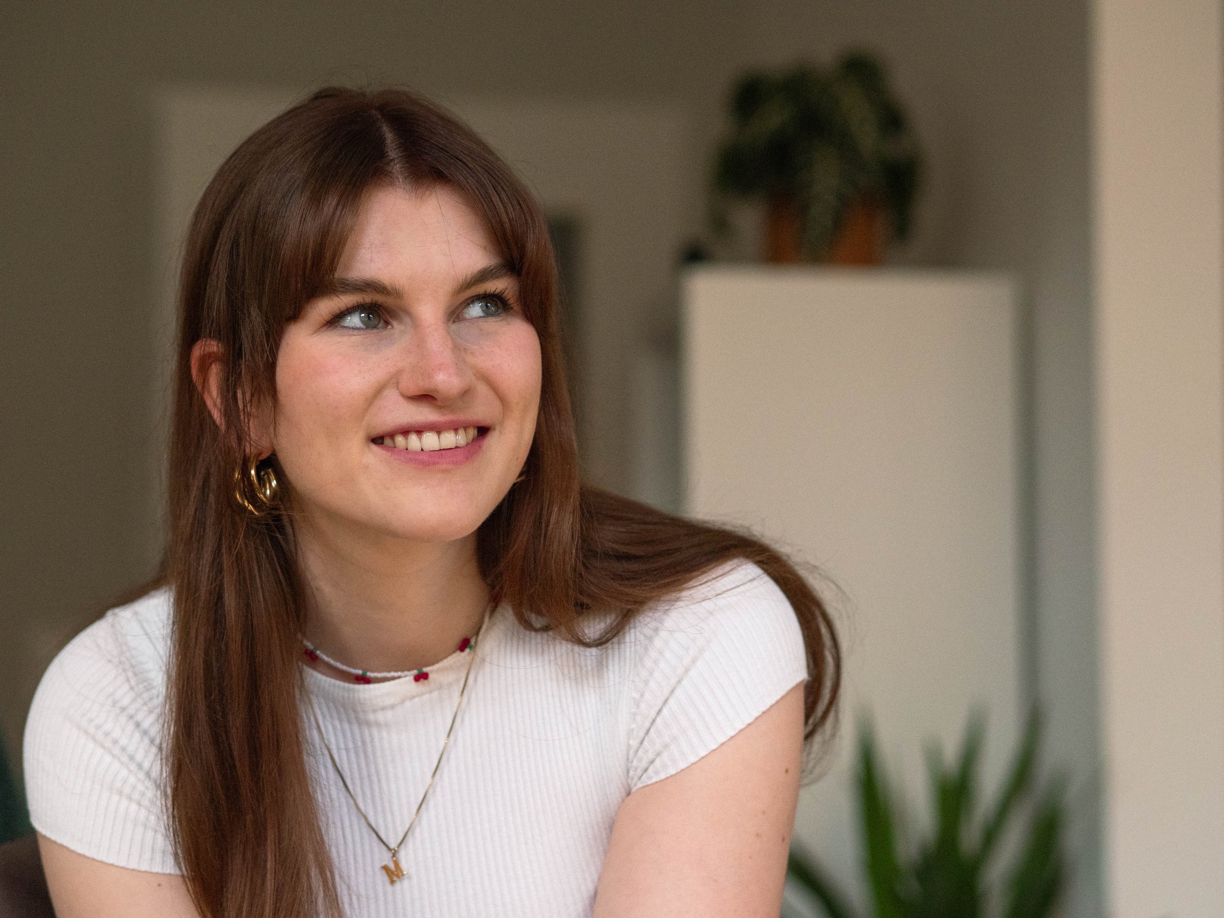 Junge Frau mit langen braunen Haaren, Oberteil aus Ribbstoff, Kette mit Anfangsbuchstabe M, lächelnd, im Innenraum mit Pflanze im Hintergrund.