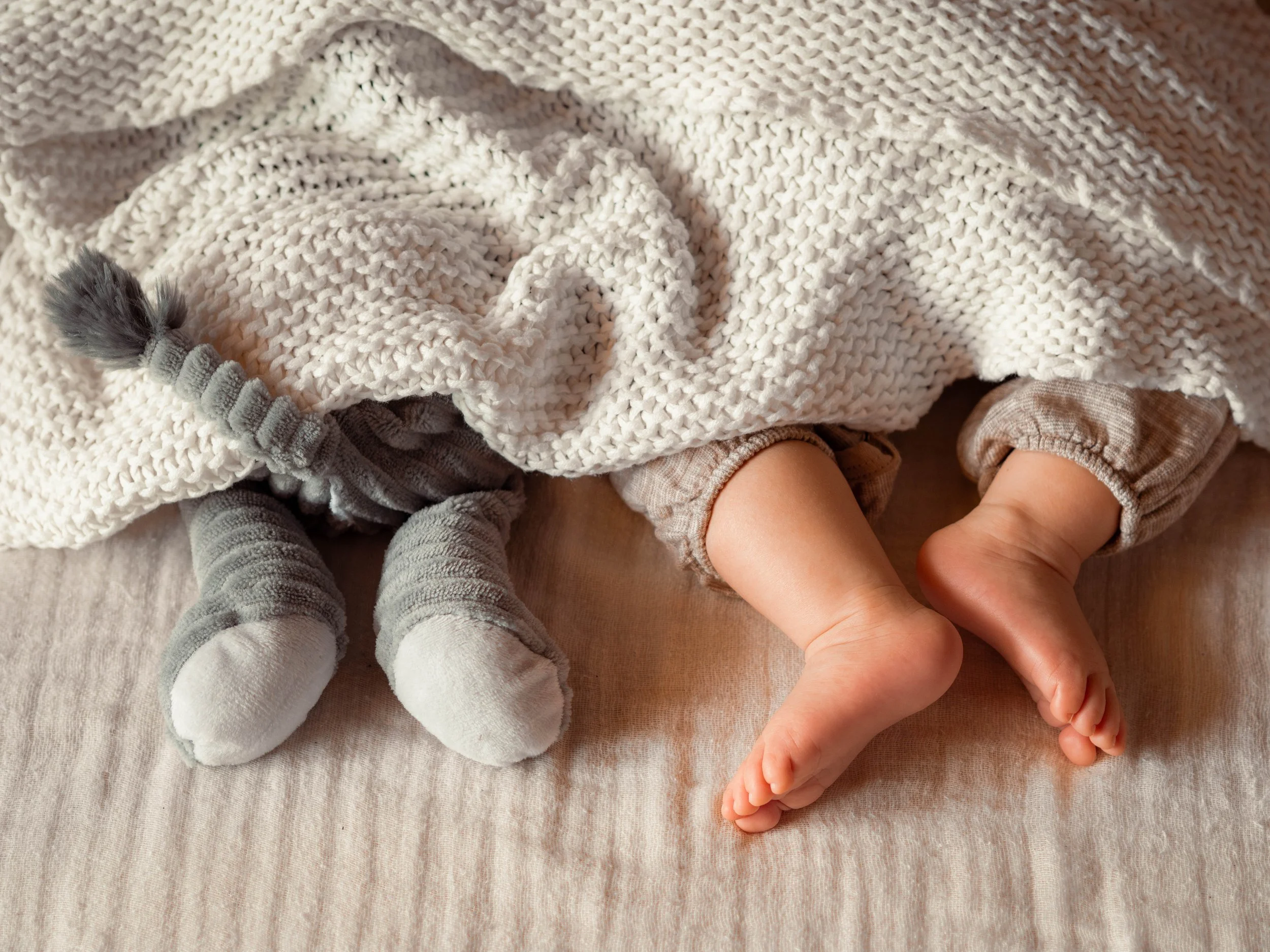 Die Fuße eines Babys und die Füße seines Kuscheltiers, wie sie nebeneinander unter einer Decke liegen.