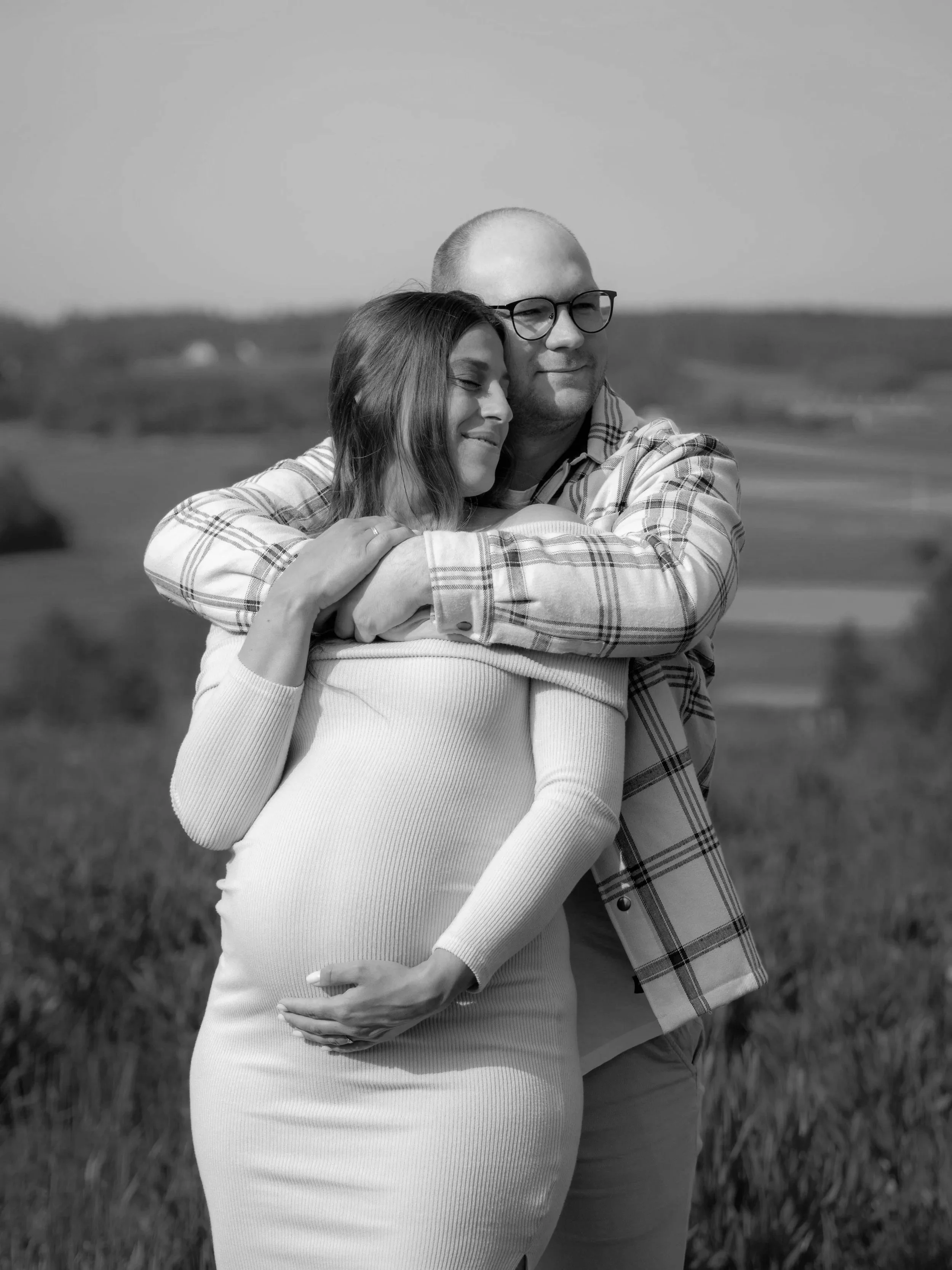 Ein Mann und eine Frau umarmen sich in einem offenen Feld, lächeln, der Mann trägt Brille und ein kariertes Hemd, die Frau trägt ein langärmeliges Kleid.