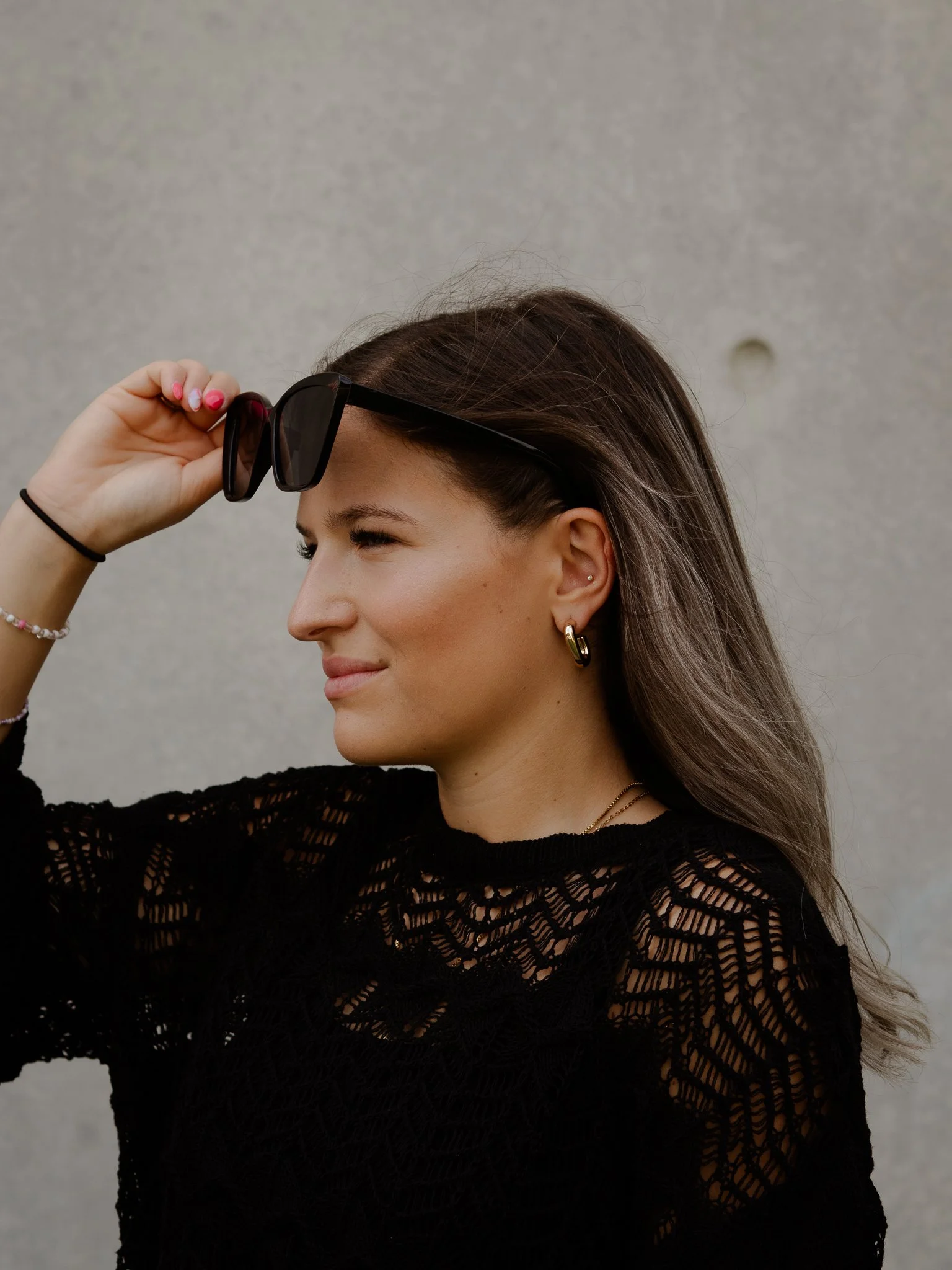Eine junge Frau, die eine schwarze Spezialbrille trägt und sie mit der rechten Hand anhebt, trägt Goldohrringe, eine Halskette und ein schwarzes, durchbrochenes Oberteil, vor einer grauen Wand.