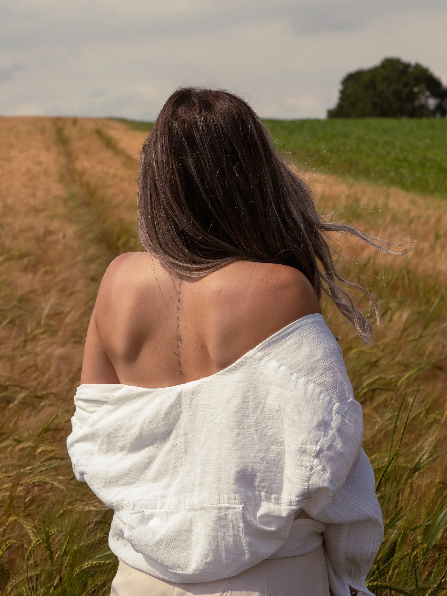 Nahaufnahme eines Schulter- und Rückenbereichs einer Frau mit braunen, lockigen Haaren, die in einem weißen, lockeren Kleid mit off-shoulder-Design steht, vor einem Feld mit Gräsern und einem Baum im Hintergrund.