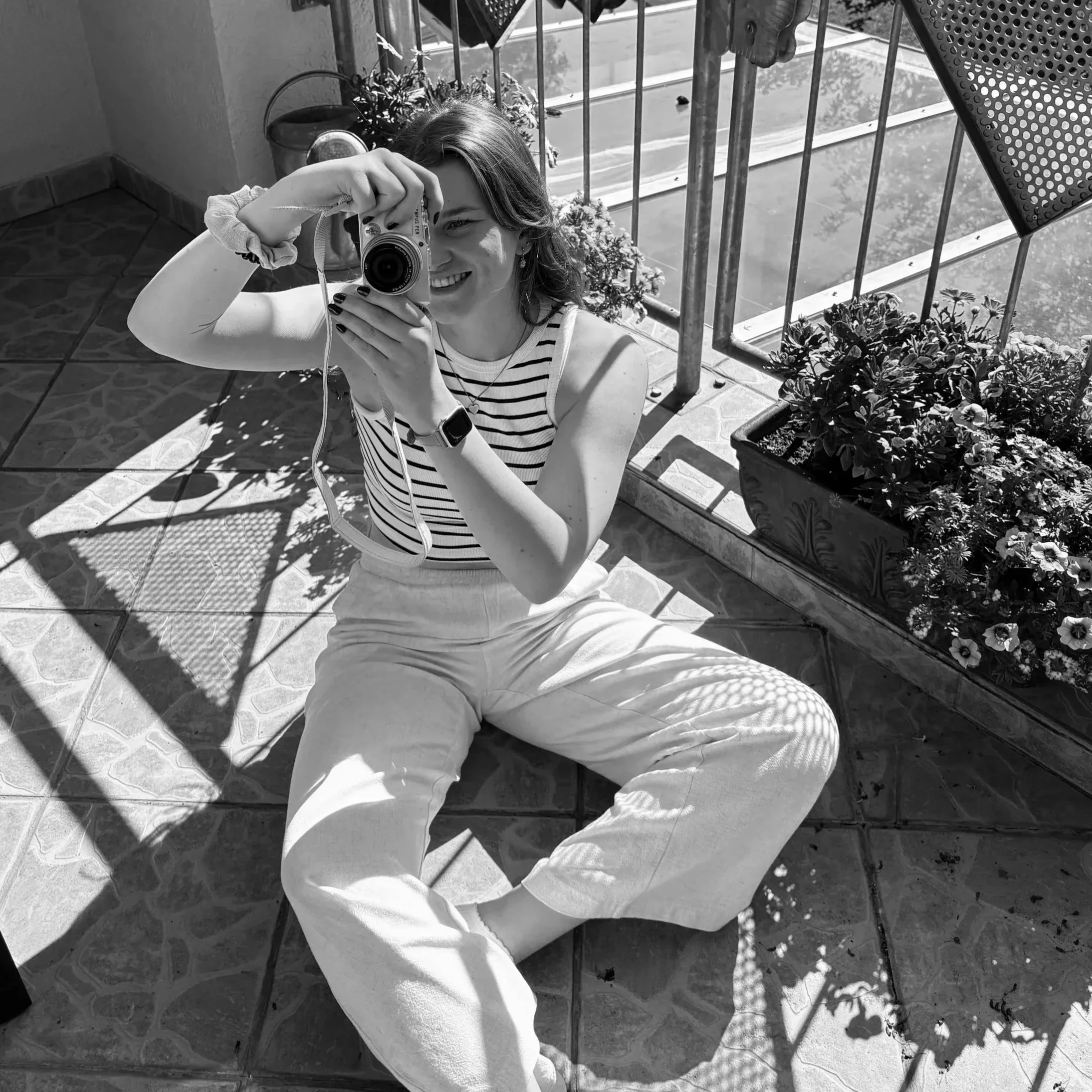 Junge Frau macht ein Selfie mit einer Kamera auf einem Balkon bei Sonnenlicht, mit Pflanzen und einem urbanen Hintergrund.