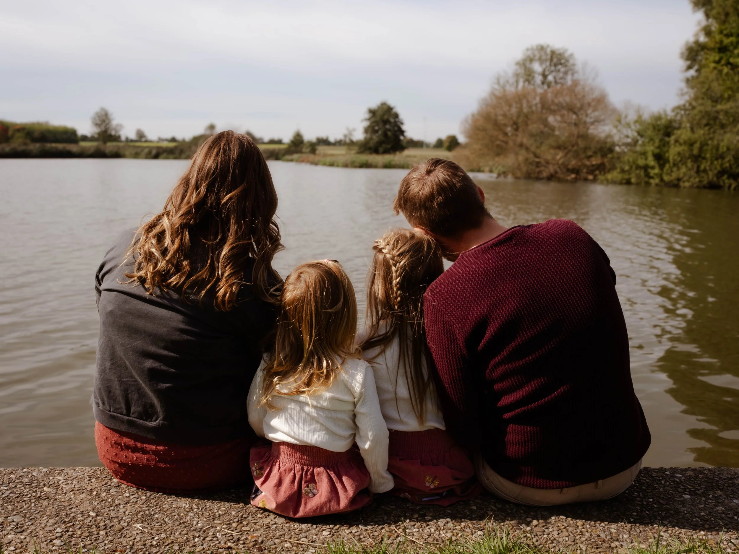 Eine Gruppe von Menschen, vermutlich Familie, sitzt am Ufer eines Flusses, blickt auf das Wasser und genießt die Natur. Es ist ein sonniger Tag im Freien.
