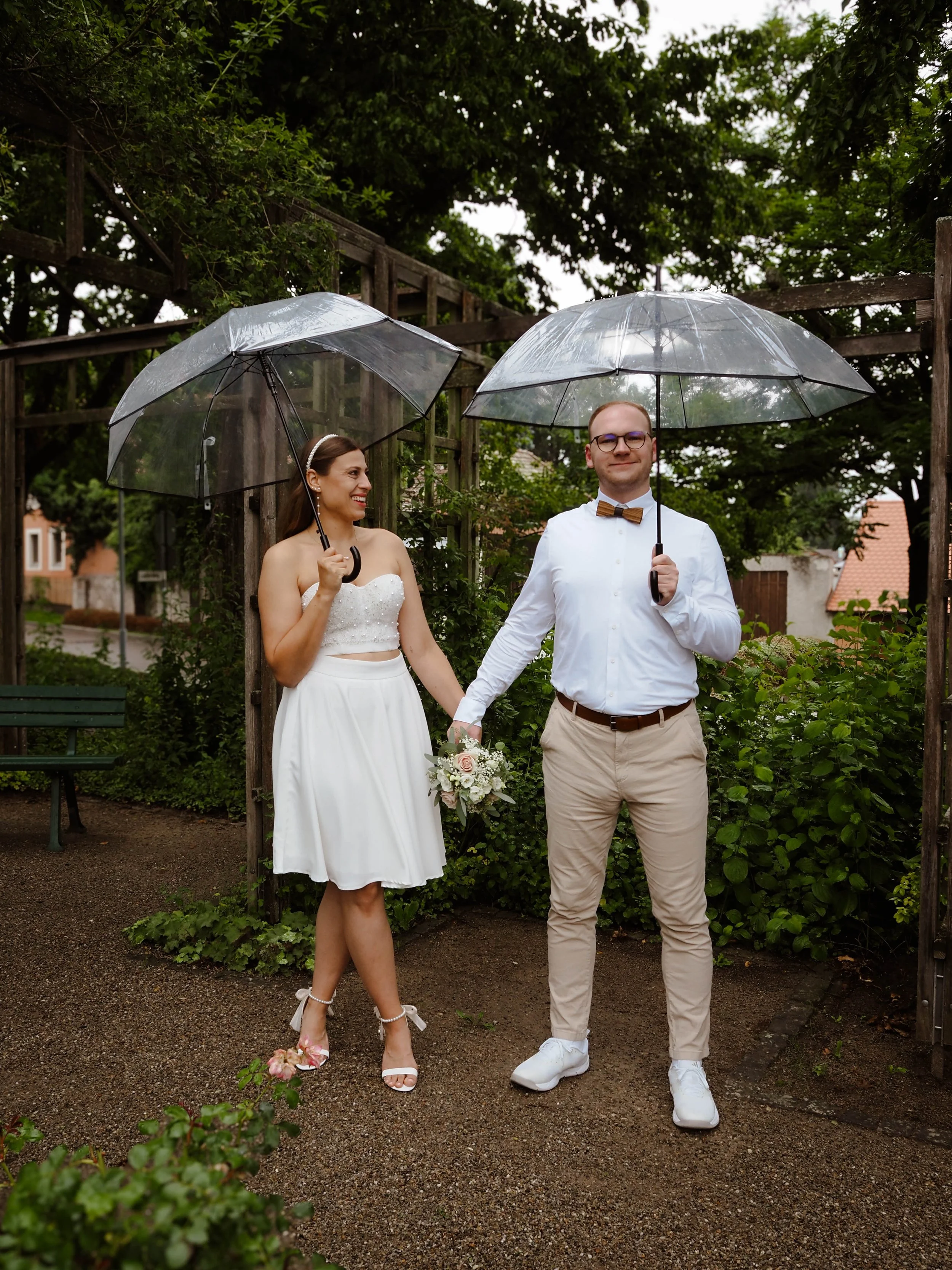 Ein brautpaar bei Regen, beide halten transparente Regenschirme, die Frau trägt ein weißes Hochzeitskleid und die Schuhe mit Schleifen, der Mann trägt ein weißes Hemd mit Fliege, sie halten sich an den Händen und stehen im Garten unter Bäumen.