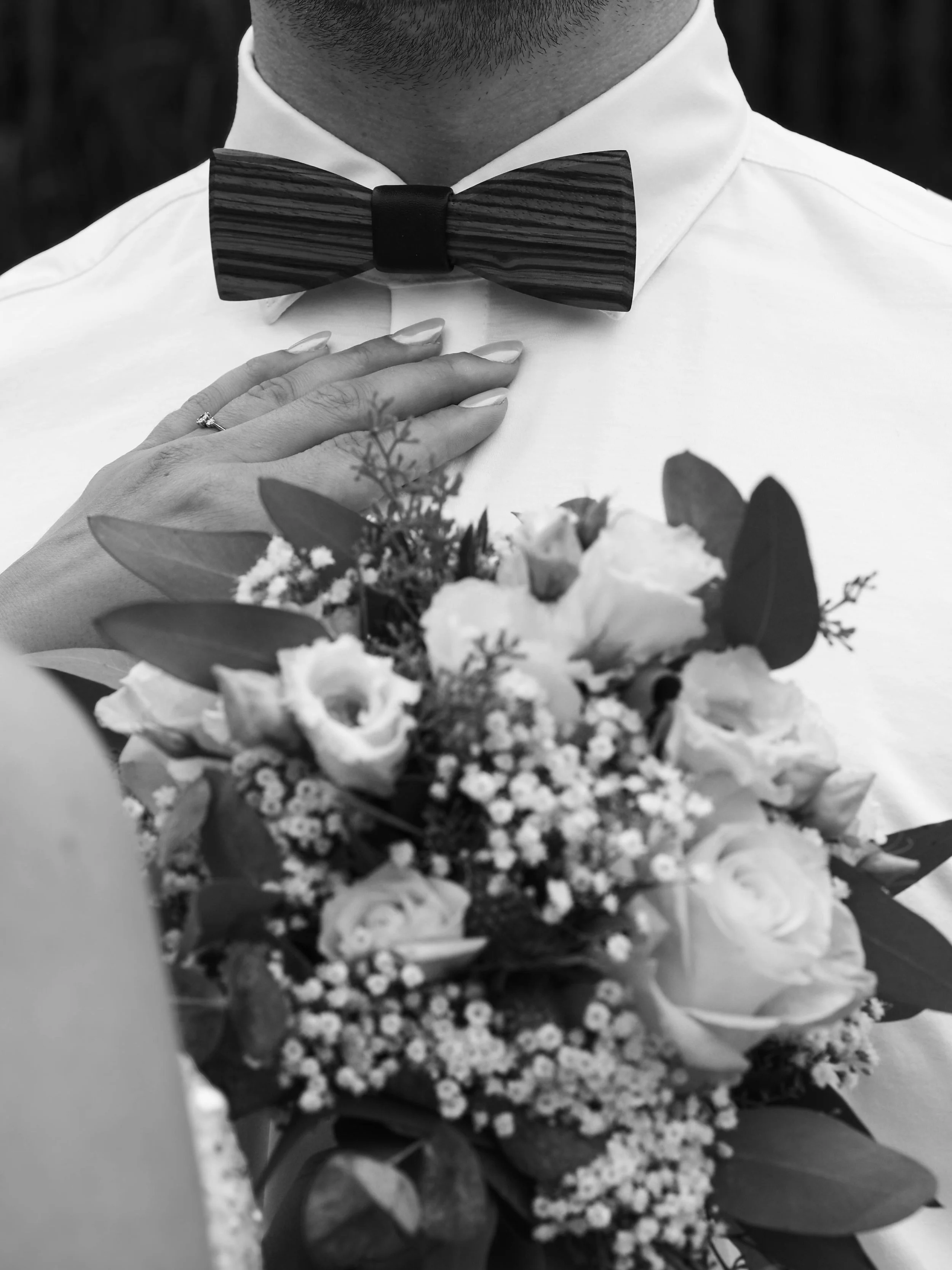Nahaufnahme eines Mannes in Weißhemd mit Fliege, der eine Hochzeitstasche mit einem Brautstrauß hält.