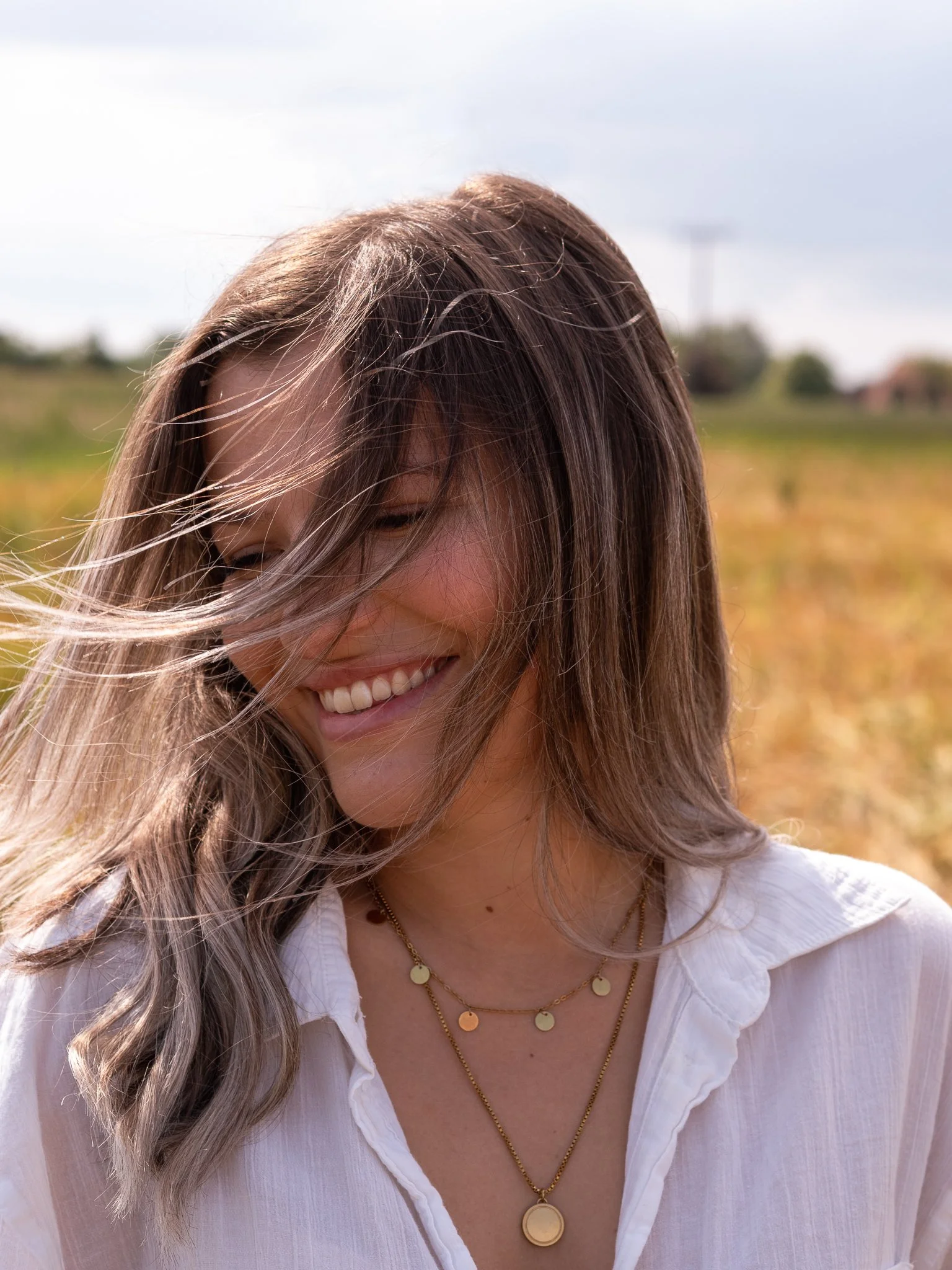Eine lachende Frau mit langen, hellbraunen Haaren, die im Freien bei sonnigem Wetter in einem Feld steht.