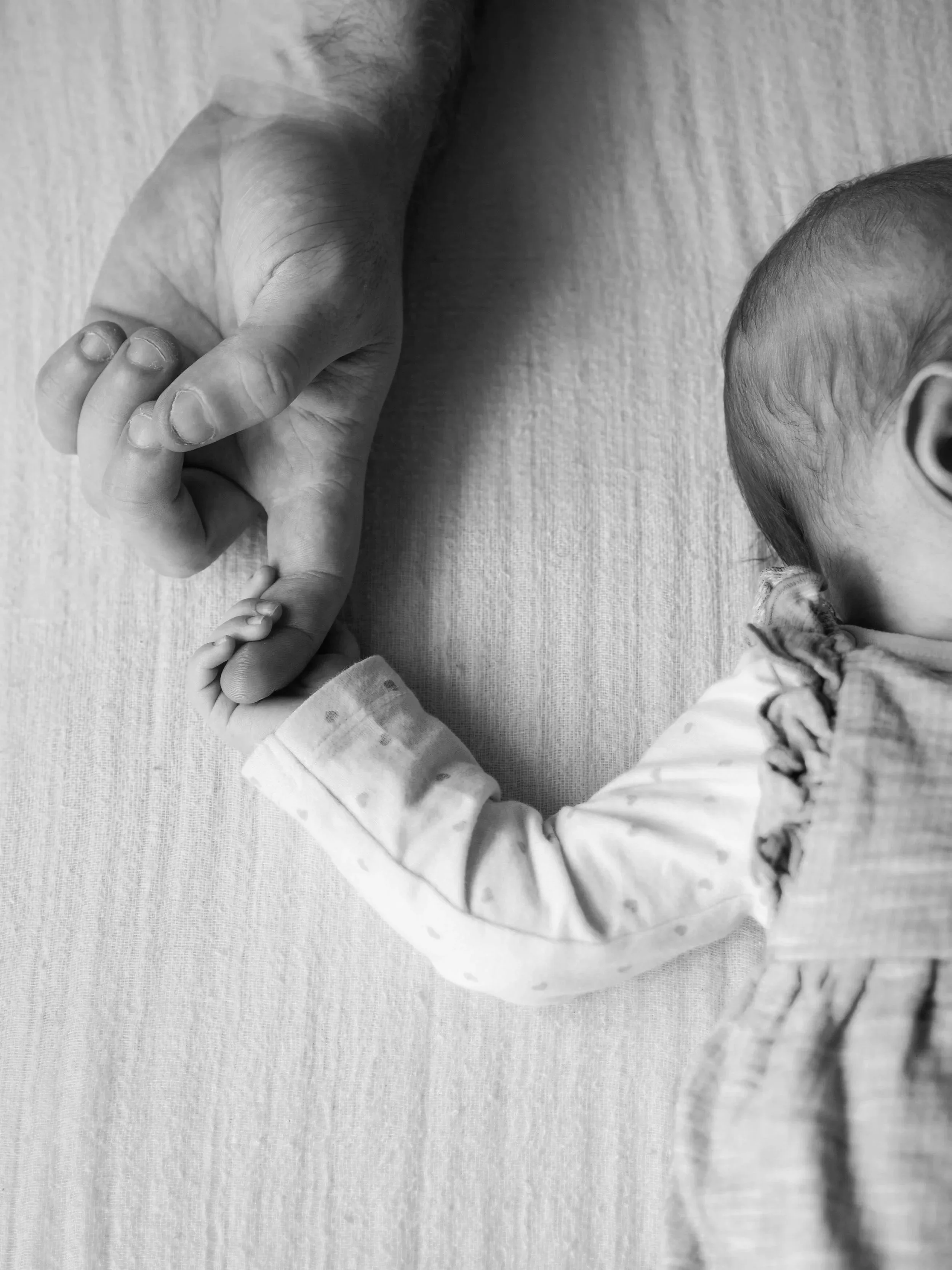 Ein Baby hält mit seiner kleinen Hand die Hand einer erwachsenen Person. Das Baby liegt auf einer weichen, hellen Unterlage.