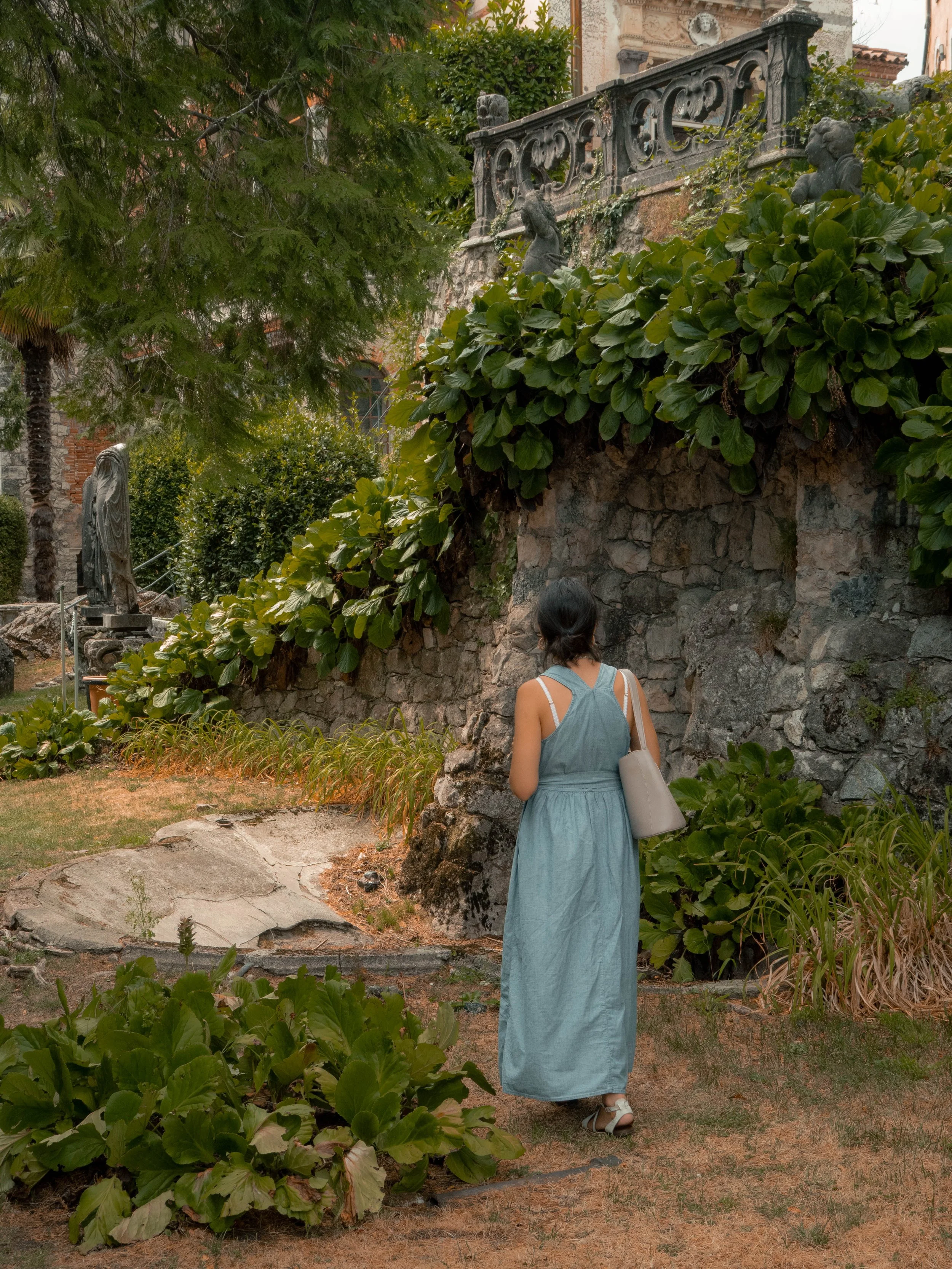Eine Frau in einem langen, blauen Kleid läuft auf einem Gartenweg. Über ihr sind grüne Pflanzen und Bäume, im Hintergrund sind antike Skulpturen und ein Geländer zu sehen.