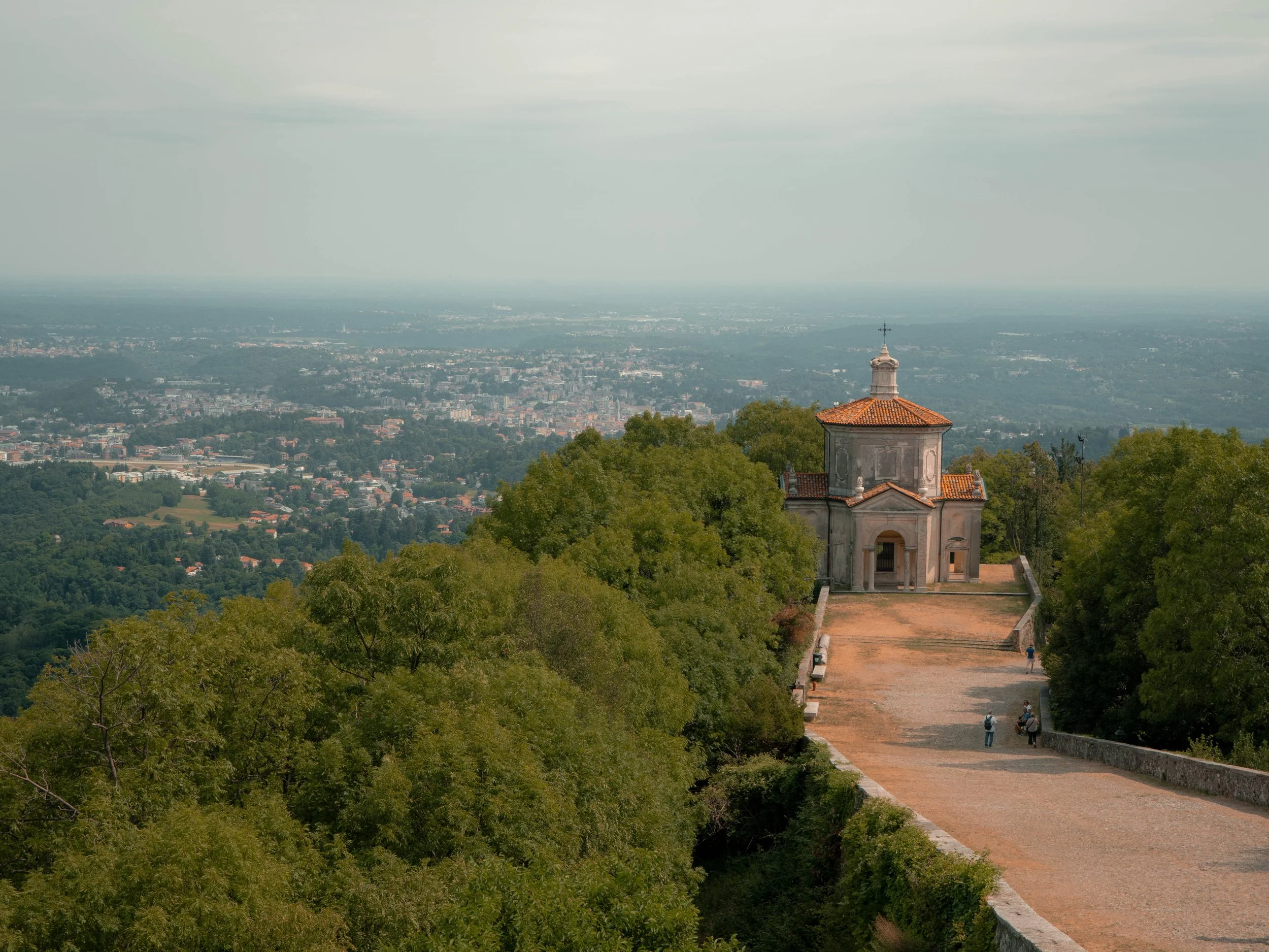 Ein serener Blick über eine Stadt, mit einem kleinen Gebäude mit roter Ziegeldach und einem Kreuz auf dem Dach, umgeben von grünen Bäumen im Vordergrund, und weite Stadtansicht im Hintergrund unter bewölktem Himmel.