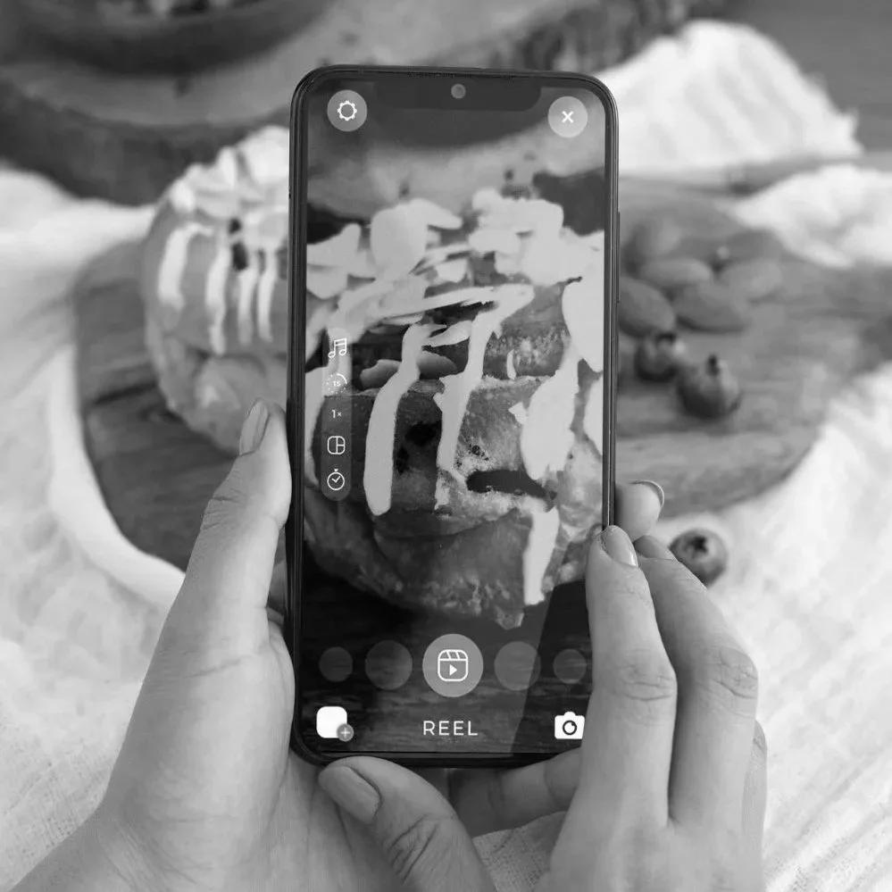 Eine Person filmt einen Kuchen mit Eis und Dekorationen auf einem Smartphone. Im Hintergrund ist der Kuchen auf einem Teller sichtbar.