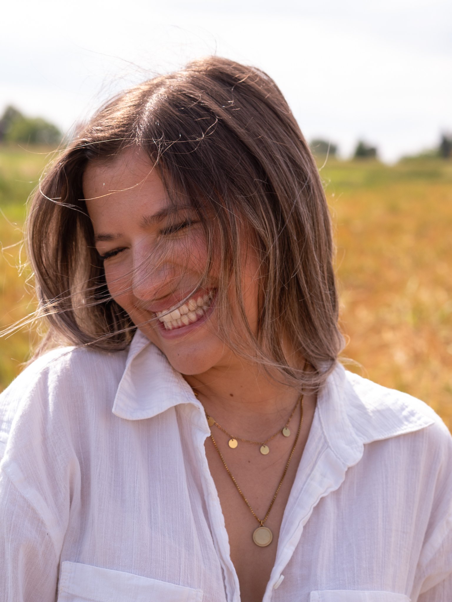 Ein lachendes Mädchen trägt eine weiße Bluse und Goldschmuck im Freien, im Hintergrund Gärten und Himmel.