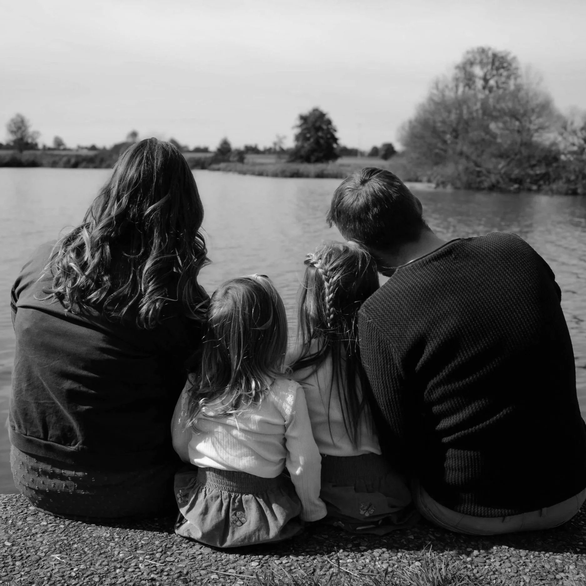 Zwei Eltern und zwei Kinder  sitzen am Ufer eines Flusses und schauen auf das Wasser, im Hintergrund Bäume unter einem bewölkten Himmel