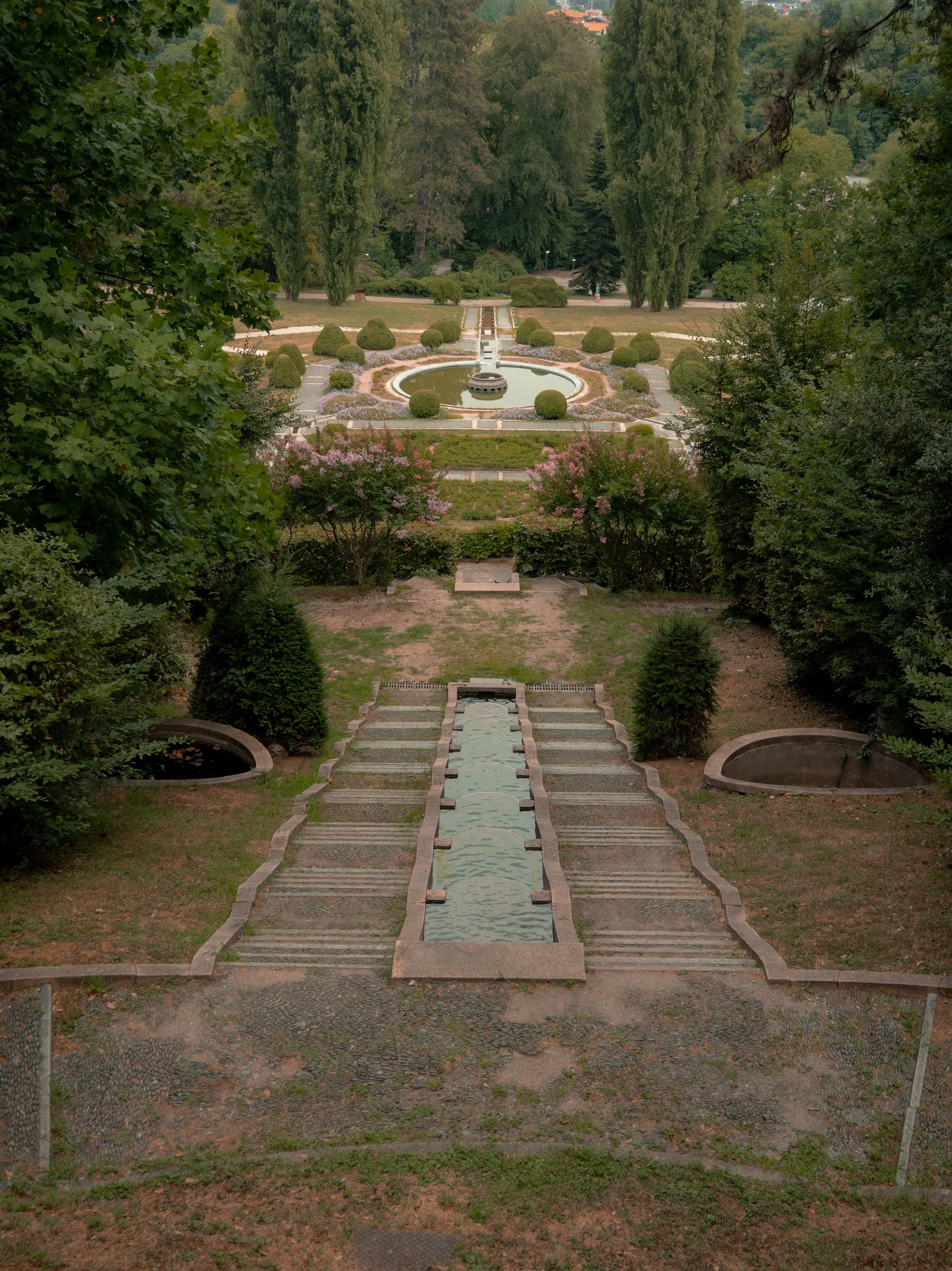 Blick auf einen gepflegten Garten mit Brunnen, Wasserweg und umgebenden Bäumen, Pflanzen und Wegen.