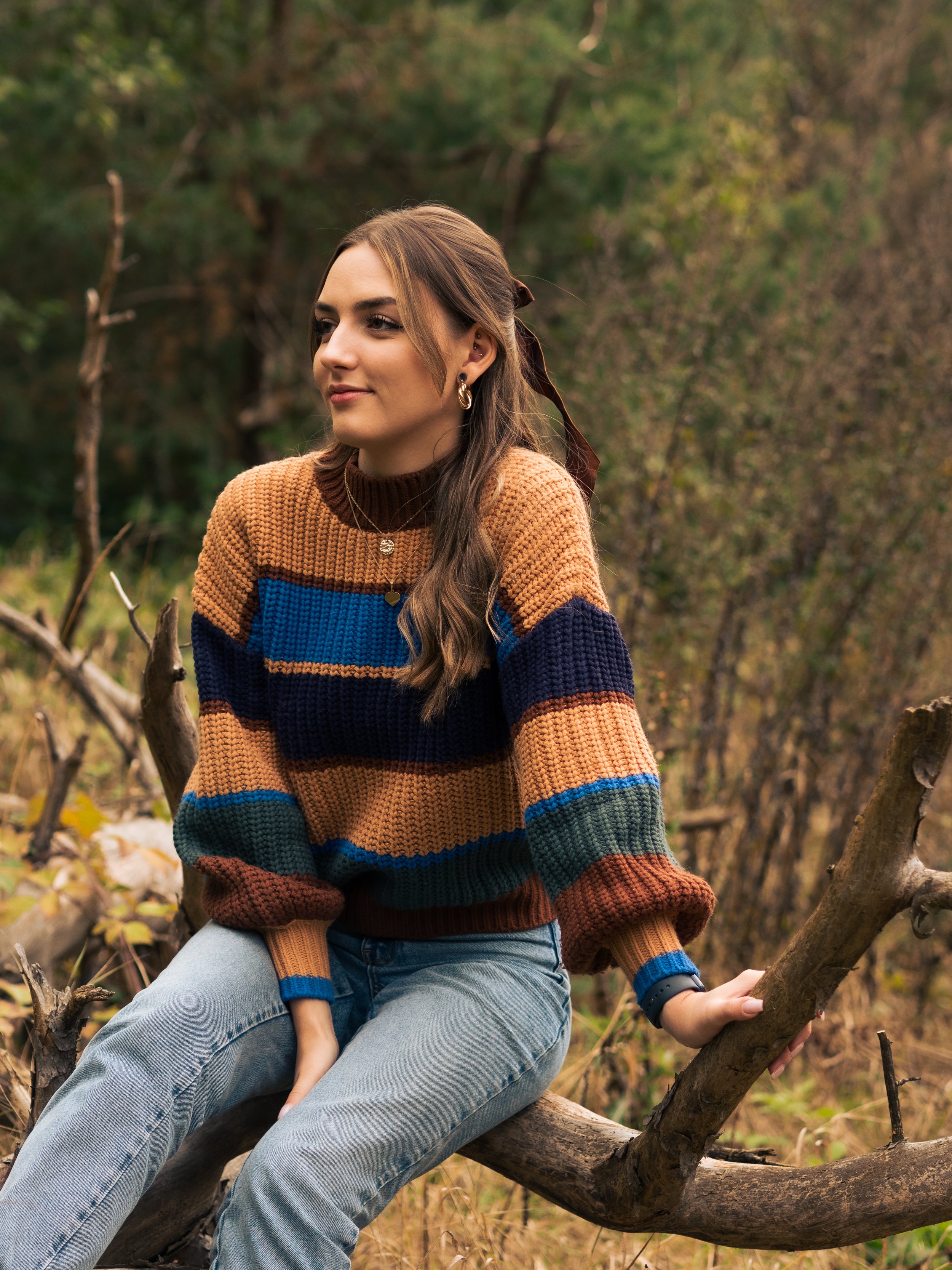 Junge Frau in farbig gestreiftem Pullover, sitzt auf Baum im Herbstwald.