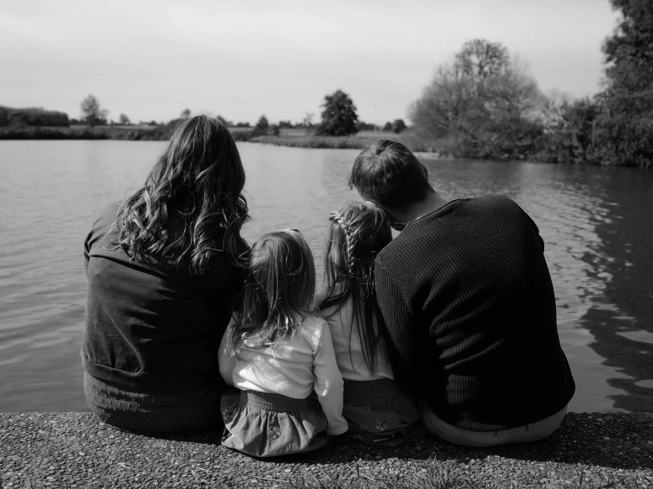 Eine Gruppe von Menschen, vermutlich Familie, sitzt am Ufer eines Flusses, blickt auf das Wasser und genießt die Natur. Es ist ein sonniger Tag im Freien.