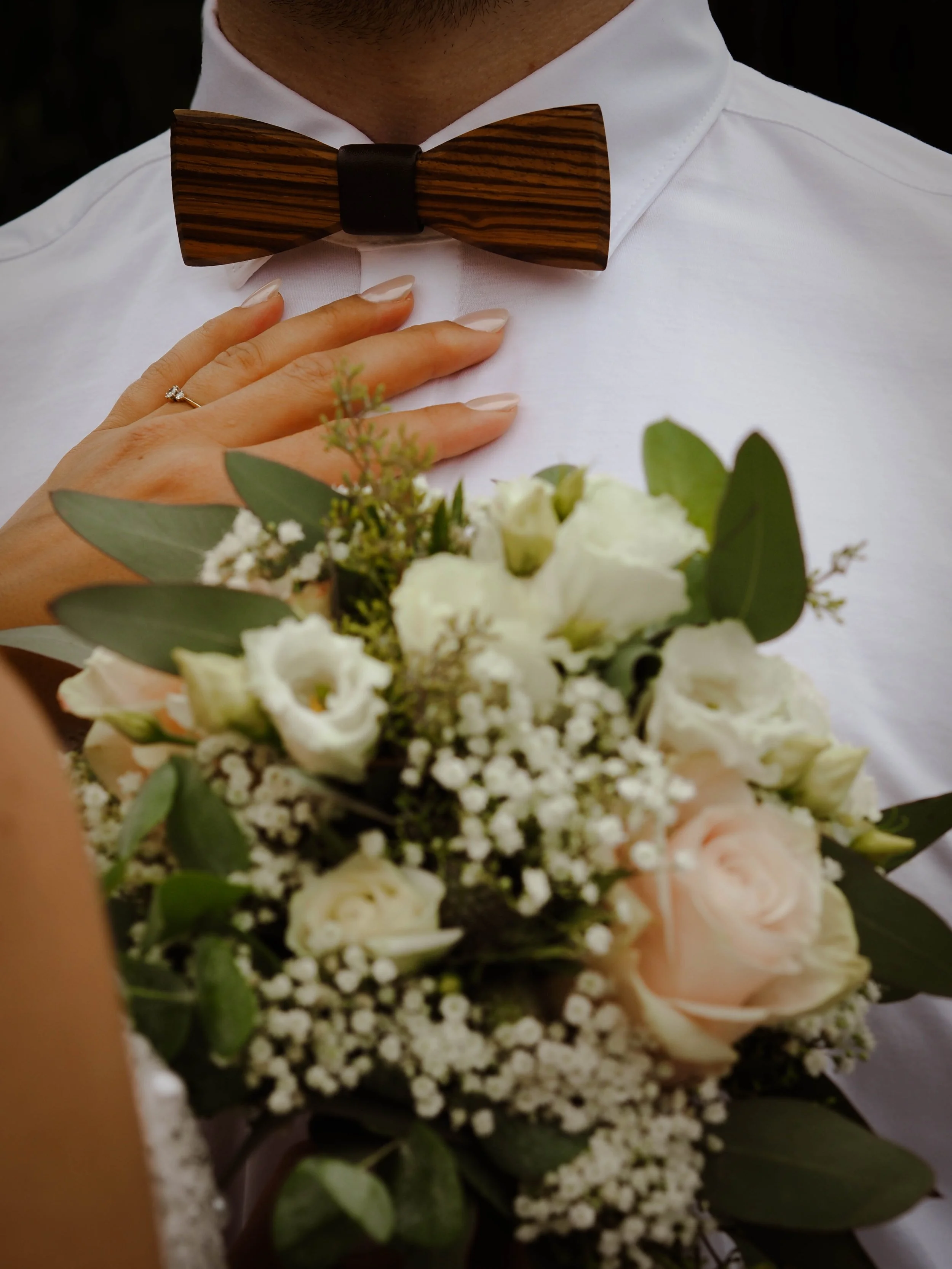 Nahaufnahme eines Mannes in weißem Hemd mit Holzmuster-Fliege, der eine Hand mit Ehering auf die Brust gelegt hält, im Vordergrund ein Blumenstrauß mit weißen und blass rosafarbenen Blumen sowie grünem Laub.