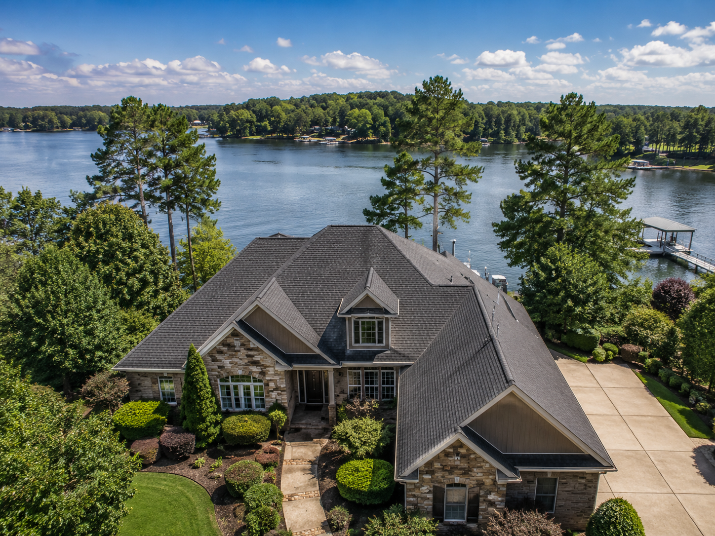 A house with a gray shingled roof and brick facade sits near a large lake with trees and a boat dock in the background, under a partly cloudy sky.