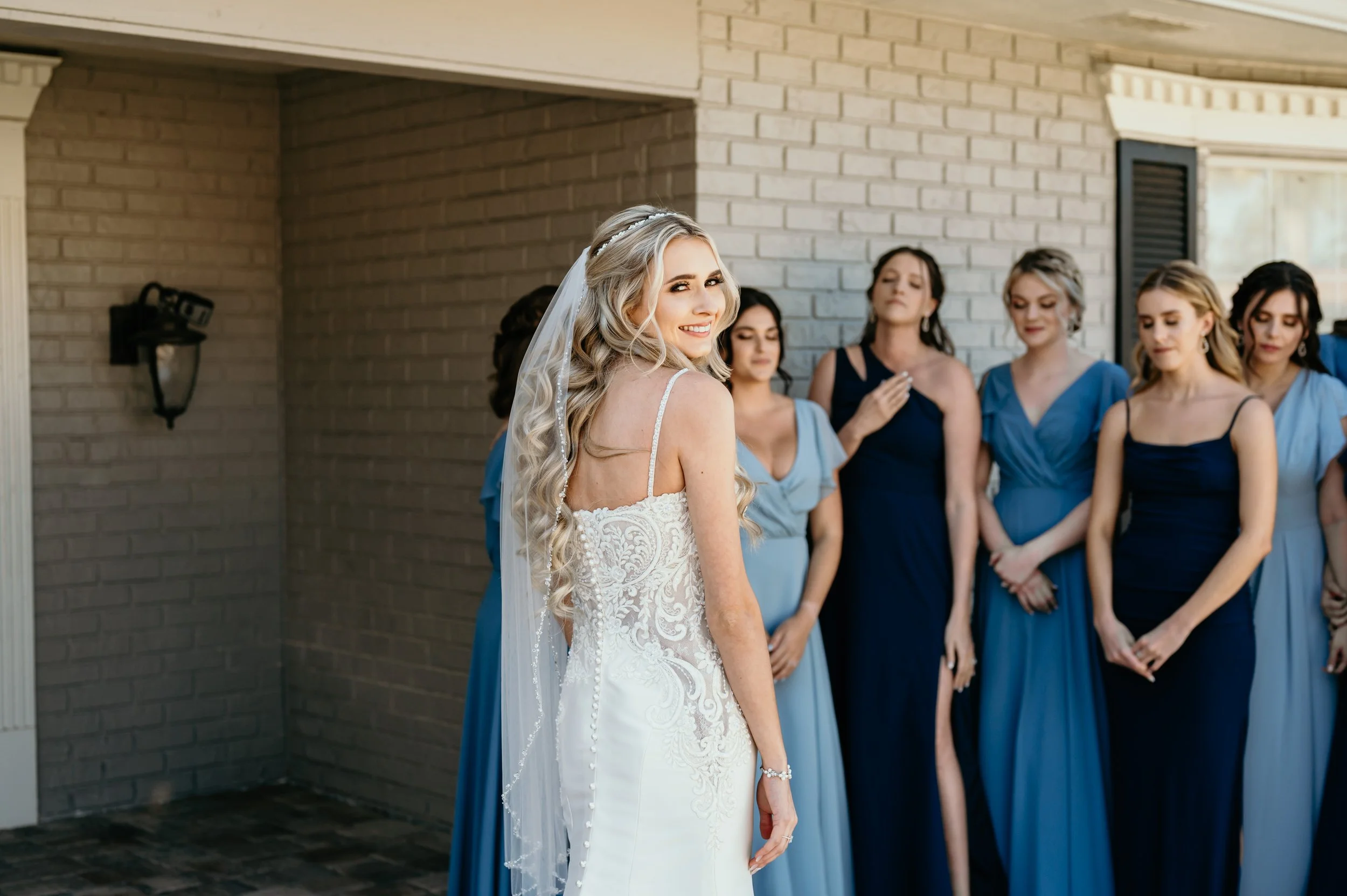 A bride in a white lace wedding dress smiling as she looks back, surrounded by bridesmaids in blue and navy dresses, standing outside near a brick house wall.