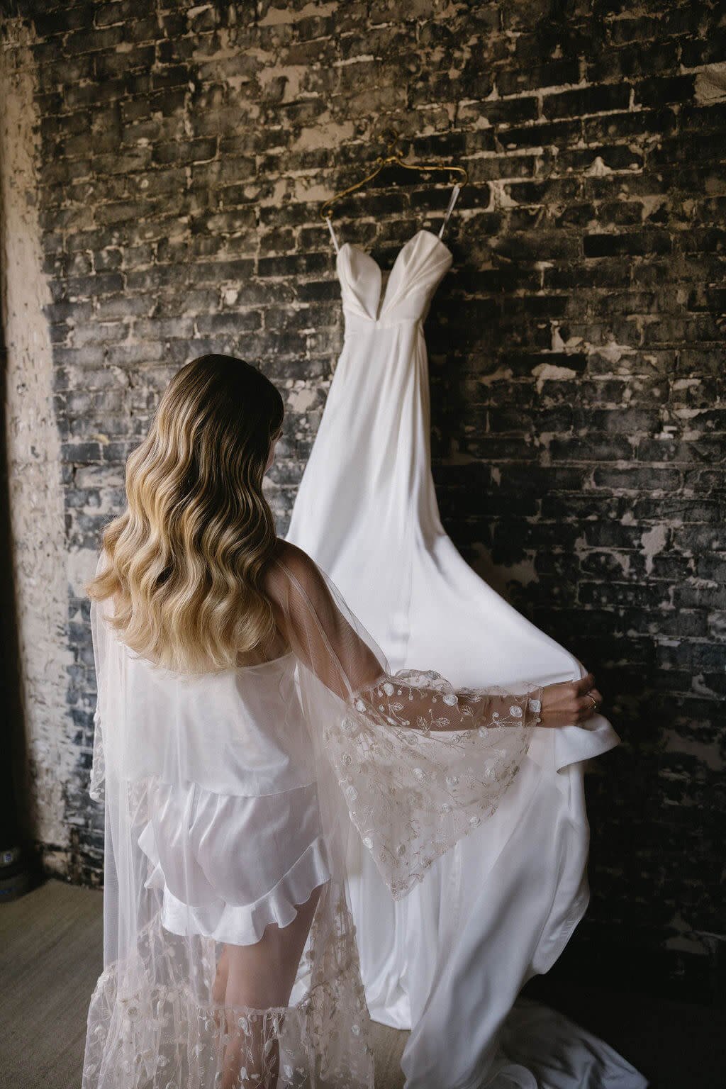 A woman with long, wavy blonde hair, wearing a sheer lace robe, looks at a white wedding dress hanging on a brick wall.