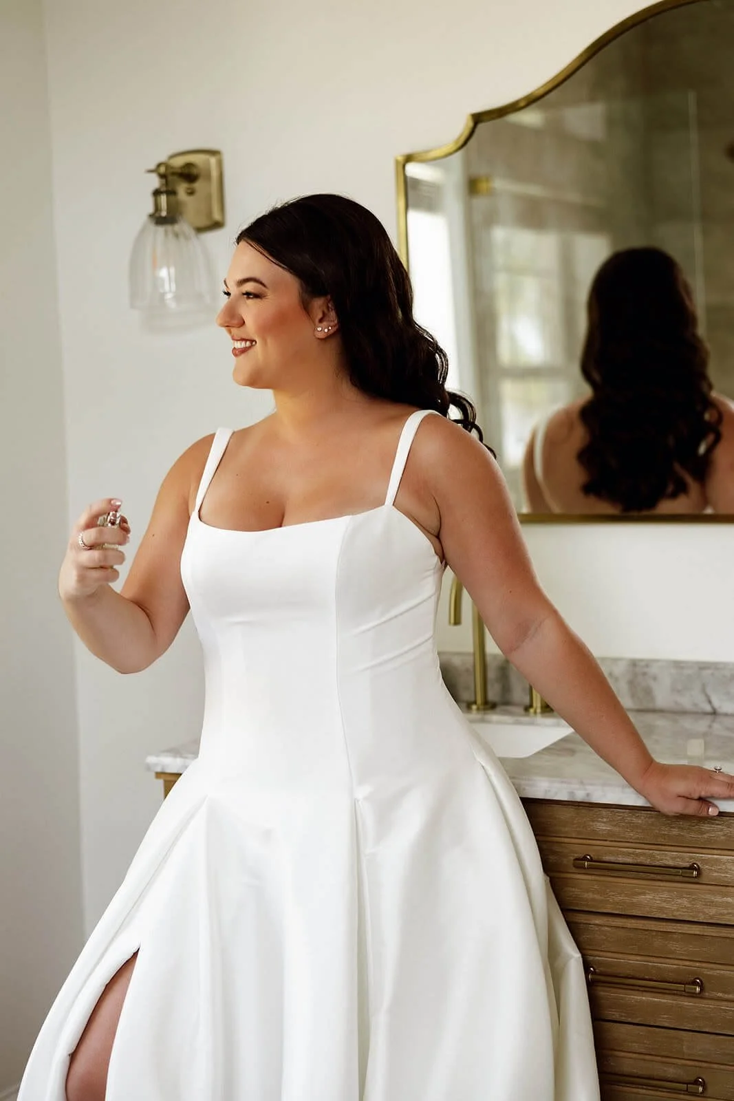 A woman in a white wedding dress spritzes perfume in front of a mirror, smiling.