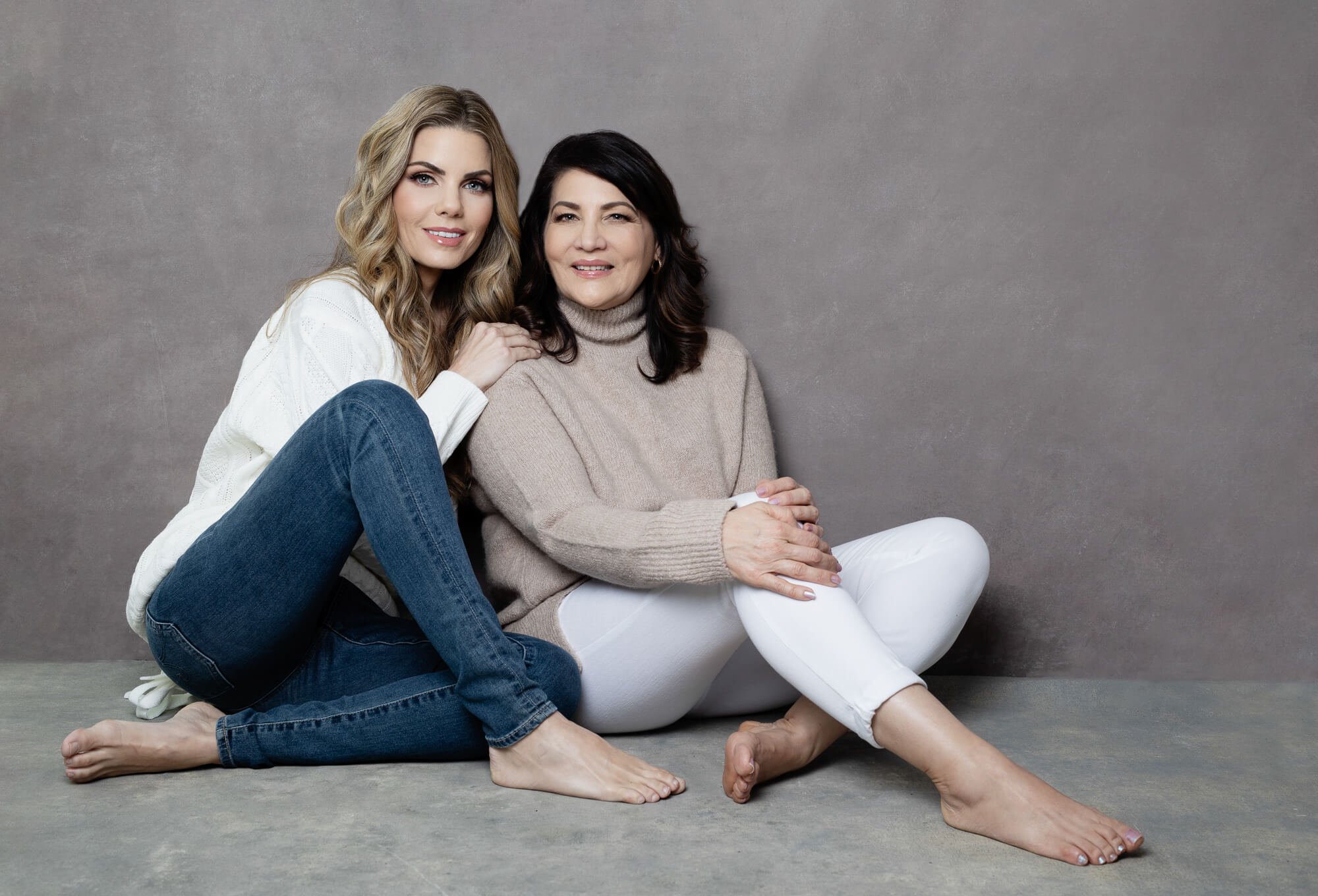 Two women sitting on the floor against a gray wall, one younger with blonde hair and the other older with dark hair, smiling and touching each other's shoulders.