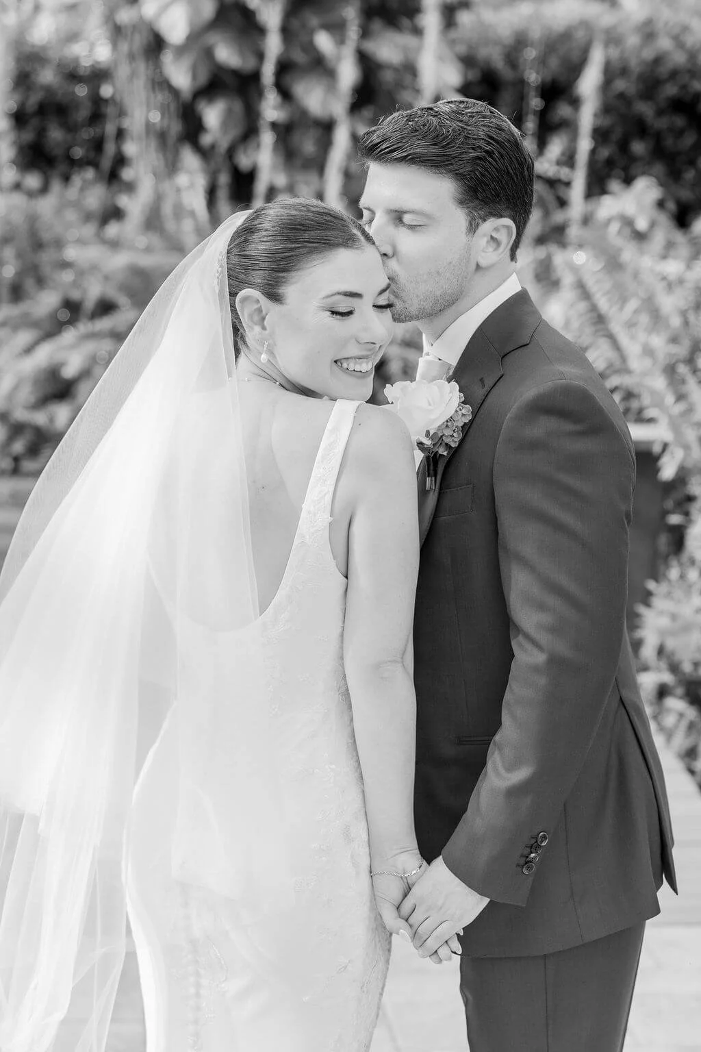 Black and white photo of a bride and groom in wedding attire, holding hands and sharing a tender moment outdoors.