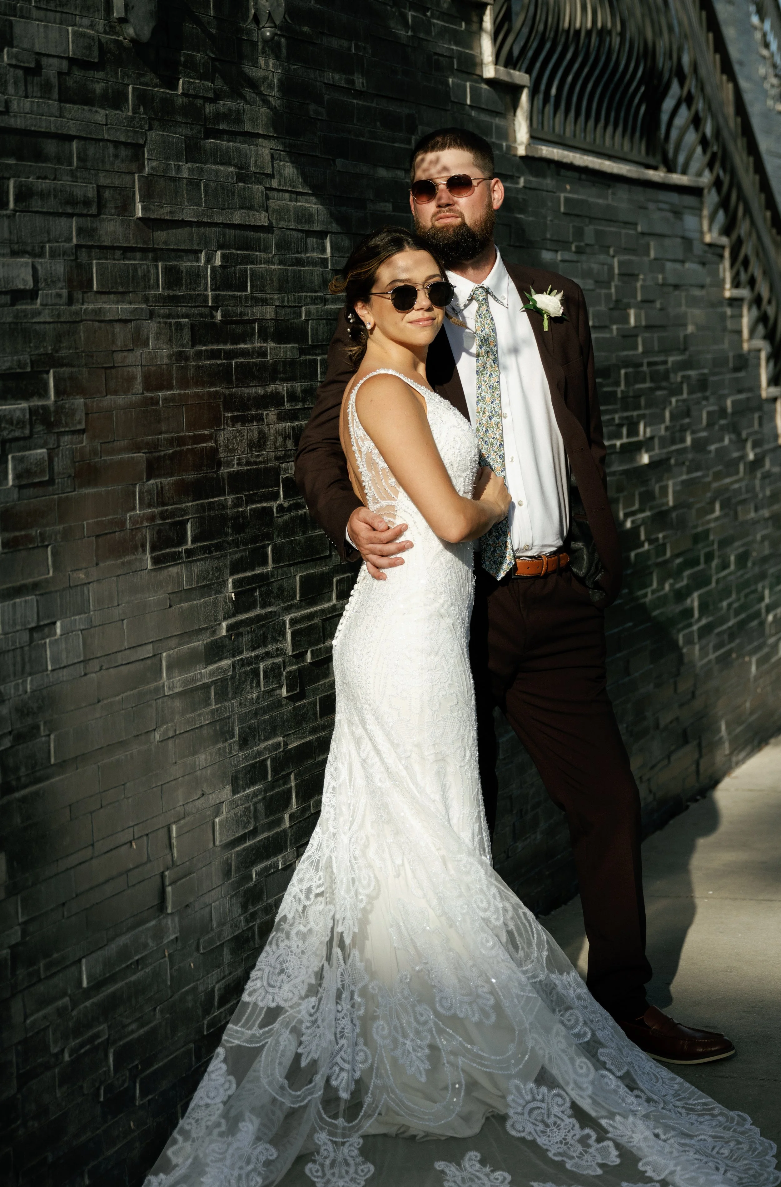 Bride in a white lace gown and sunglasses leaning against a groom in a dark suit and glasses, standing next to a black brick wall on a sunny day.