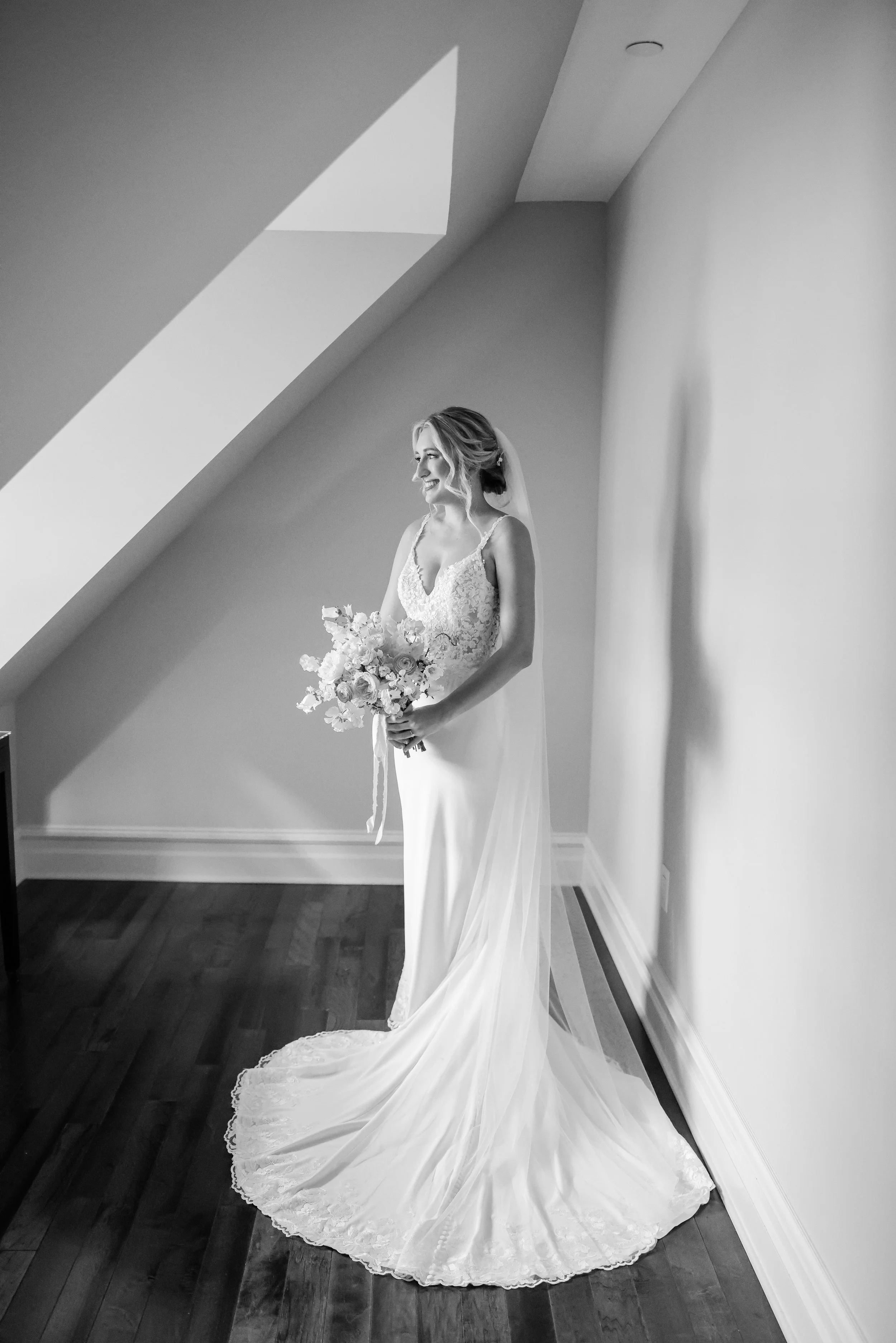 Black and white photo of a bride standing indoors, holding a bouquet, wearing a lace wedding dress with a long train, looking to the side, near a sloped ceiling with a skylight.