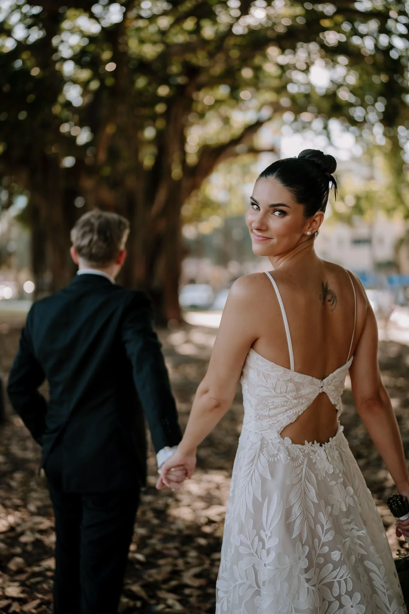 A bride and groom holding hands outdoors in a park setting, with the only the bride facing the camera smiling surrounded by trees.