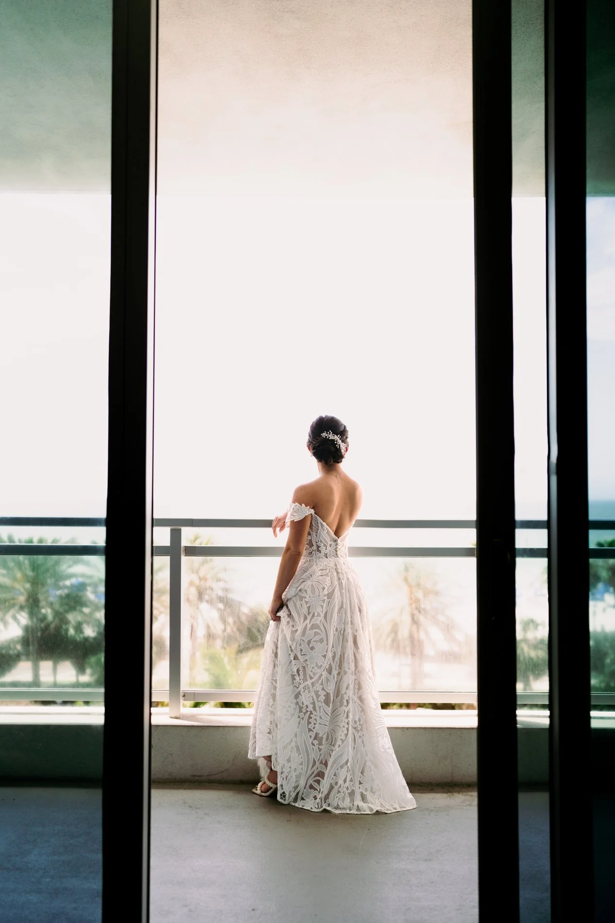 A woman in a white wedding dress standing on a balcony, facing away, looking out at a bright sky and distant trees.
