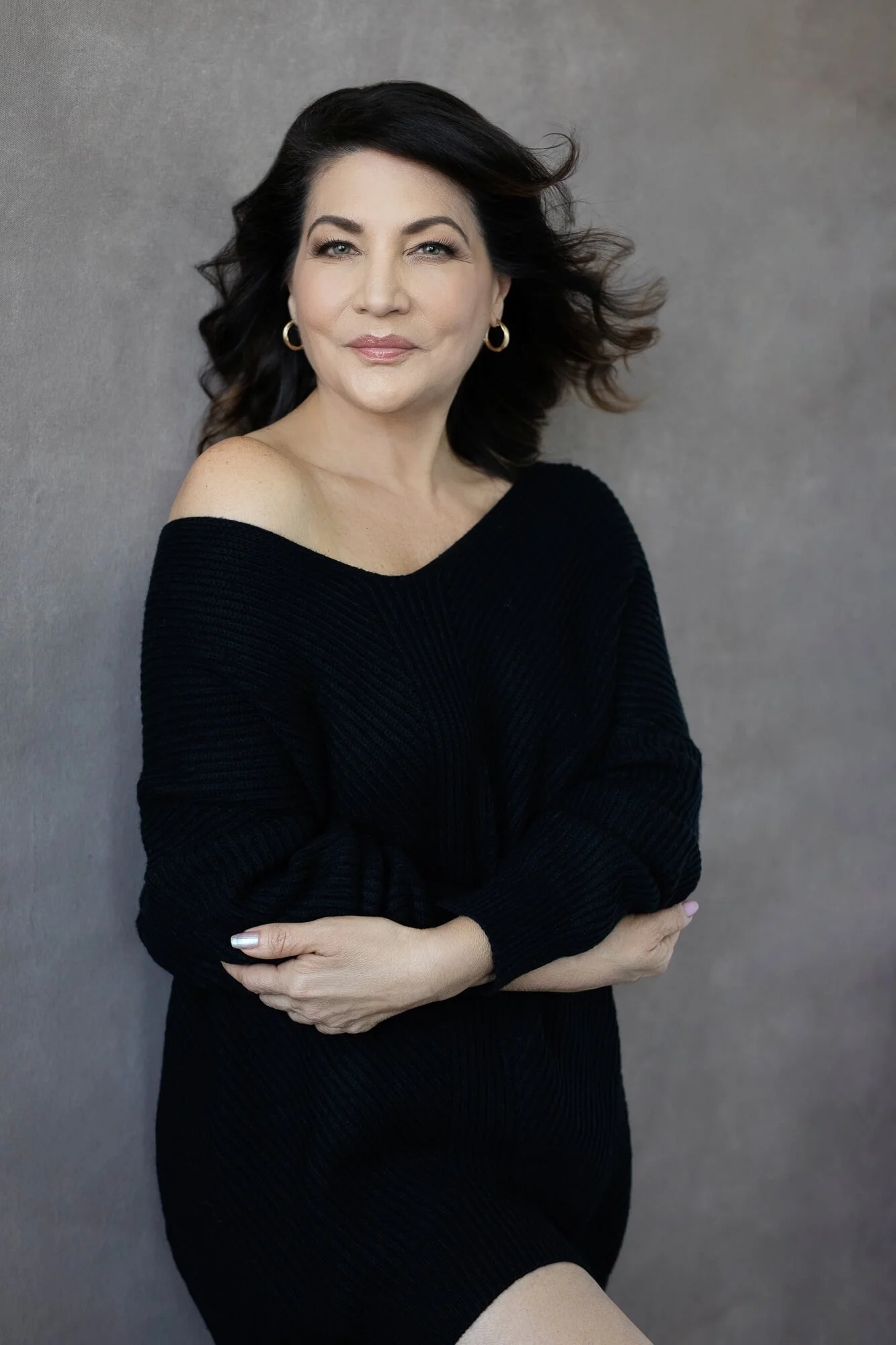 A woman with dark hair wearing a black off-the-shoulder sweater, standing against a gray background and smiling.