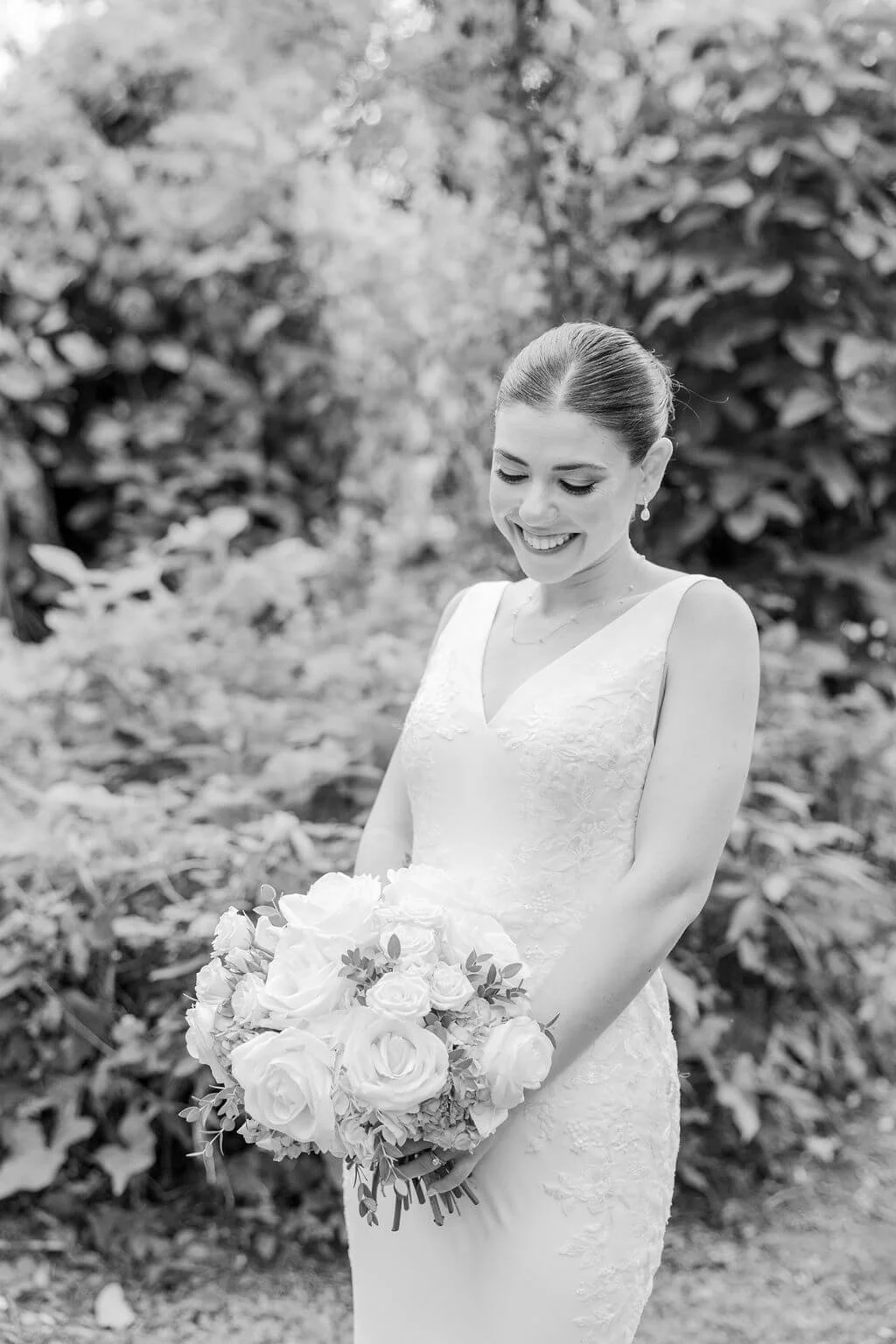 Black and white photo of a smiling bride holding a bouquet of roses, outdoors with trees and foliage in the background.