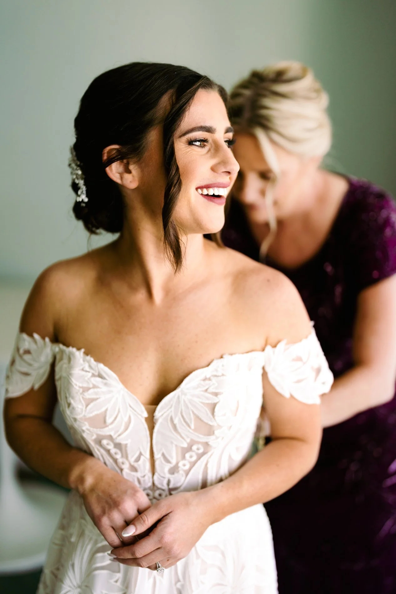 A woman in a white wedding dress smiling as another woman helps her get ready.