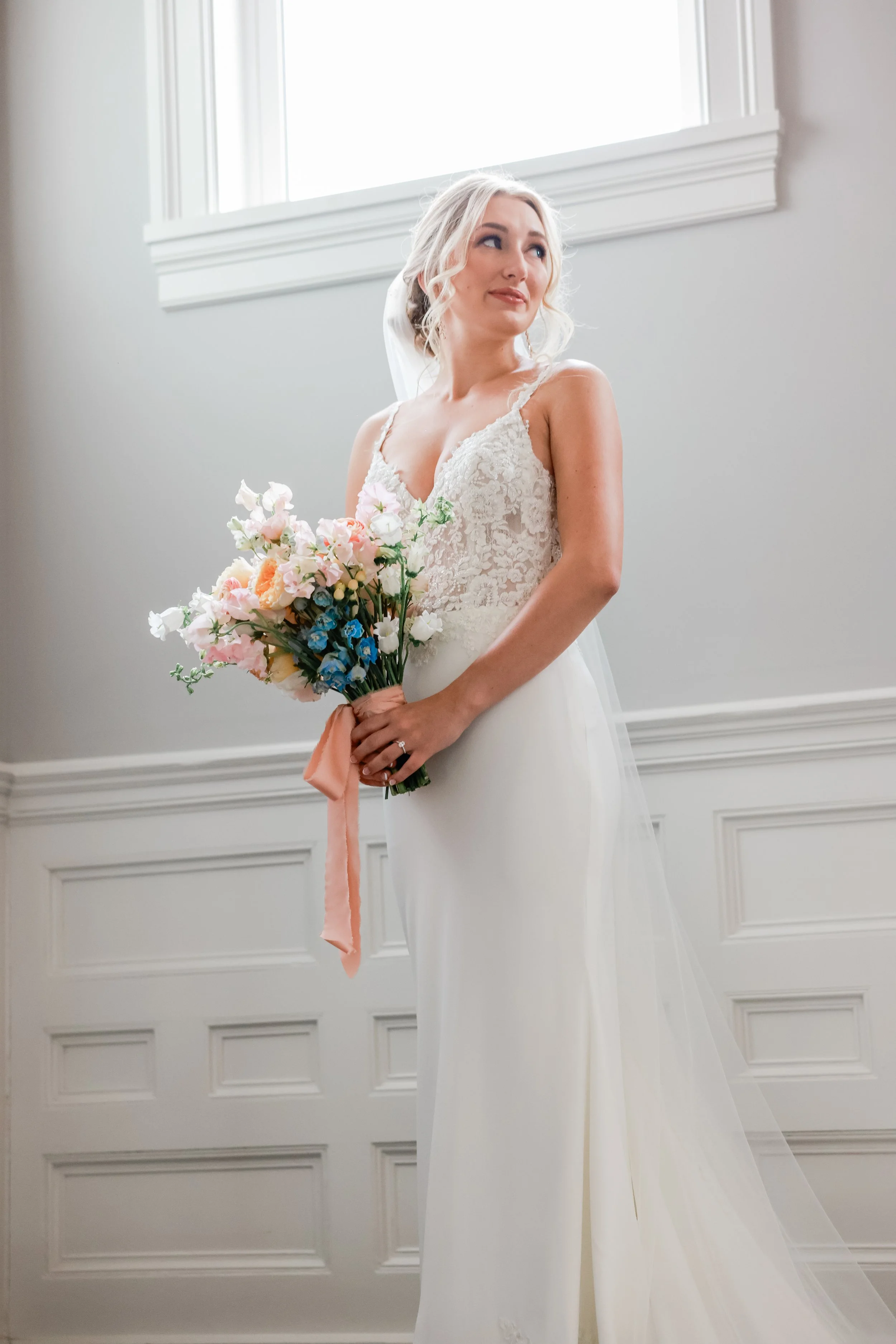 A bride in a white lace wedding dress holding a bouquet of pink, white, and blue flowers, standing near a window with white wall paneling in the background.