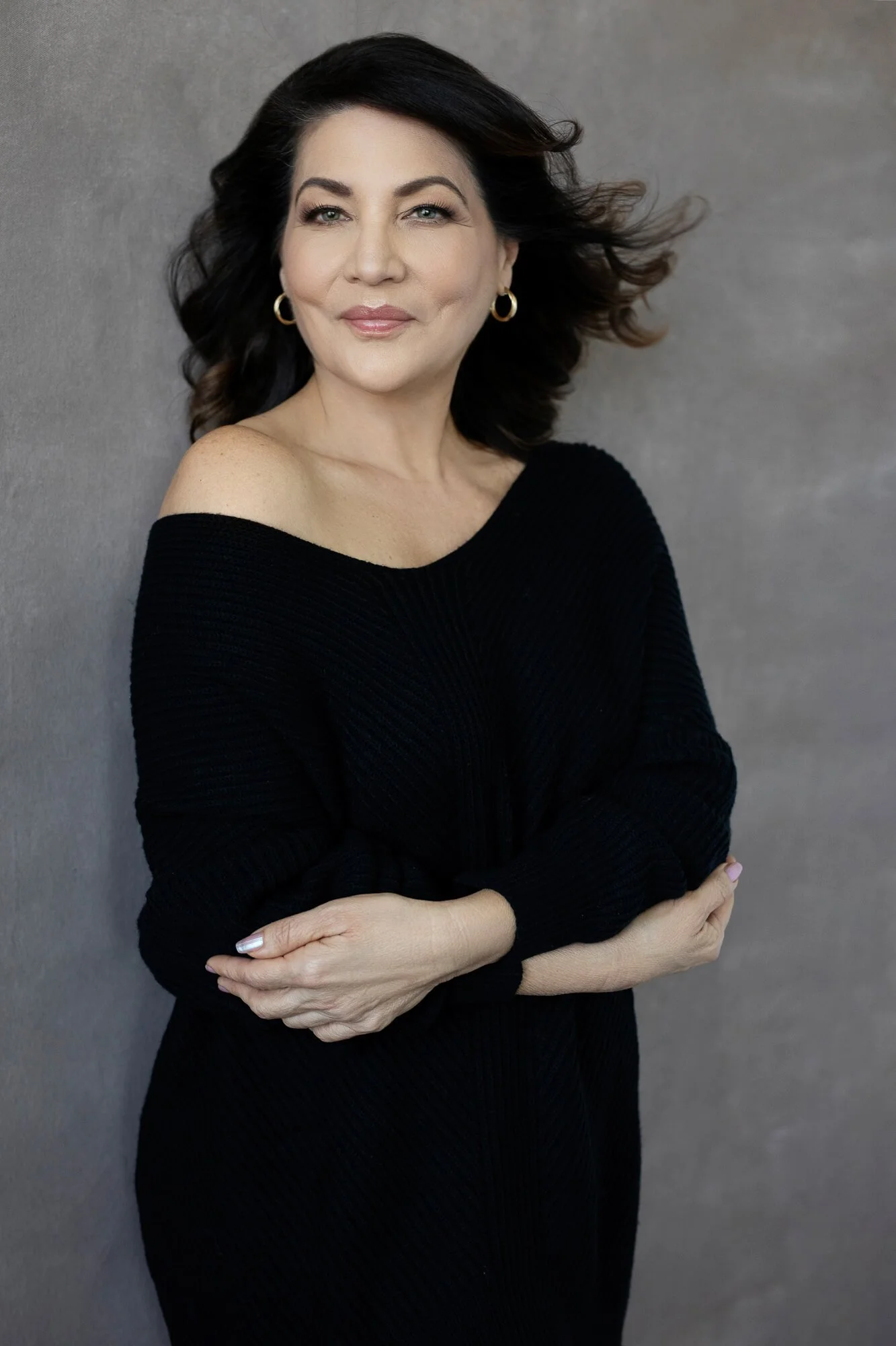 A woman with dark hair and fair skin wearing a black off-shoulder sweater, standing against a gray background, looking at the camera.