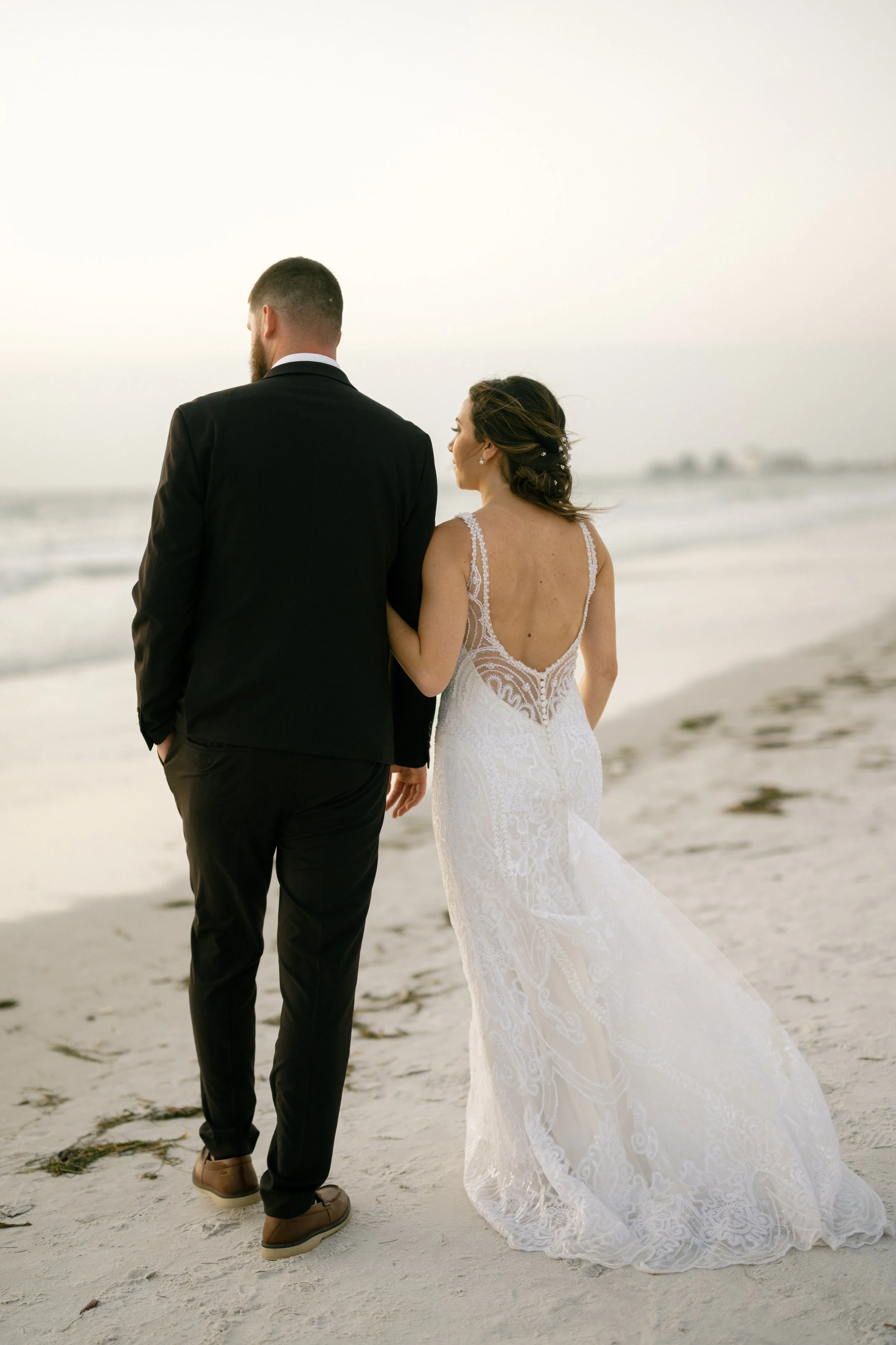 A bride and groom walk hand in hand along the beach at sunset, with the ocean and shoreline in the background.