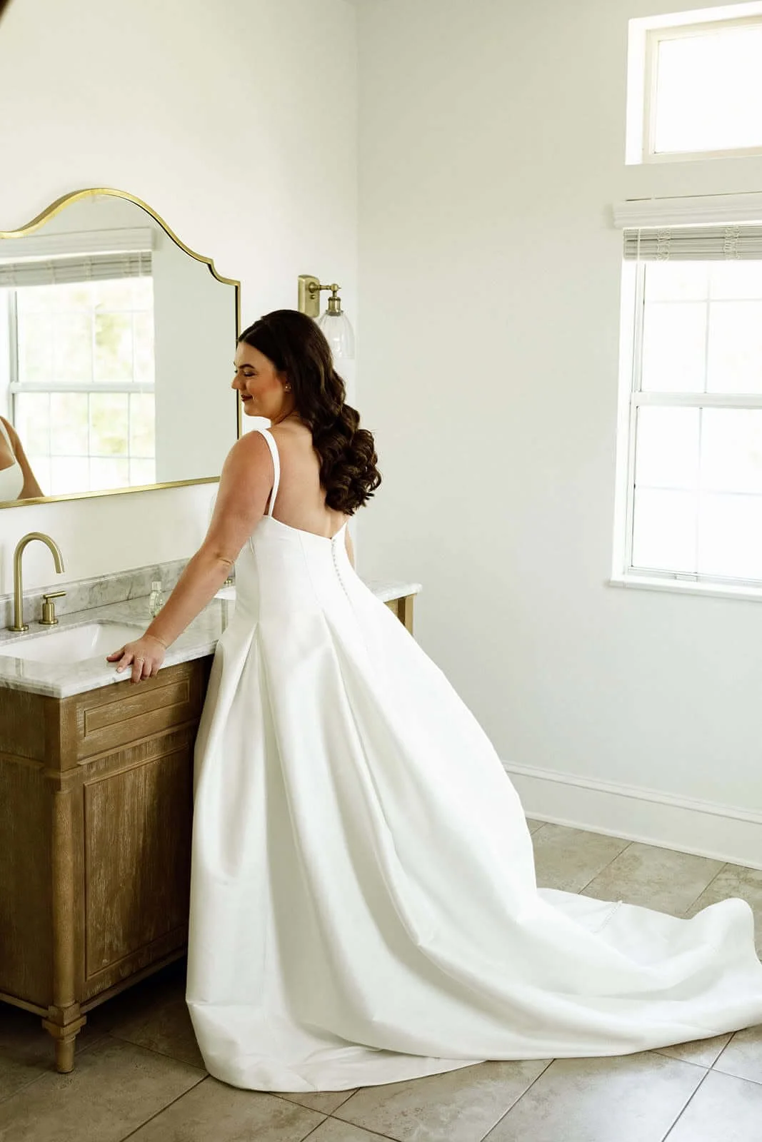 Woman in a white wedding dress standing by a bathroom sink in a bright room with large windows.