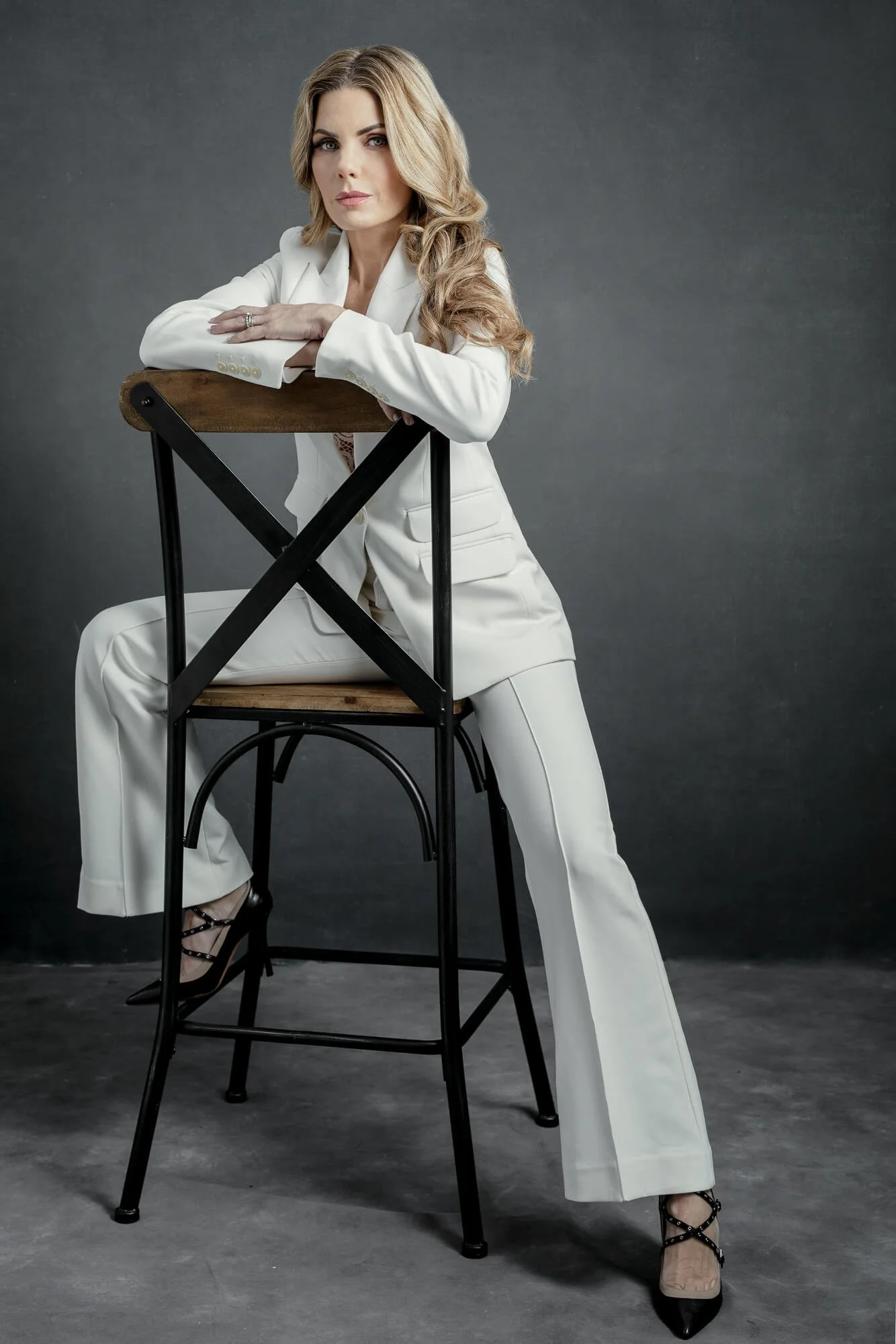 A woman in a white suit sitting on a tall wooden and metal chair, leaning forward with her arms crossed on the chair's back, with long blonde hair, makeup, and black strappy high heels, posed against a dark gray background.