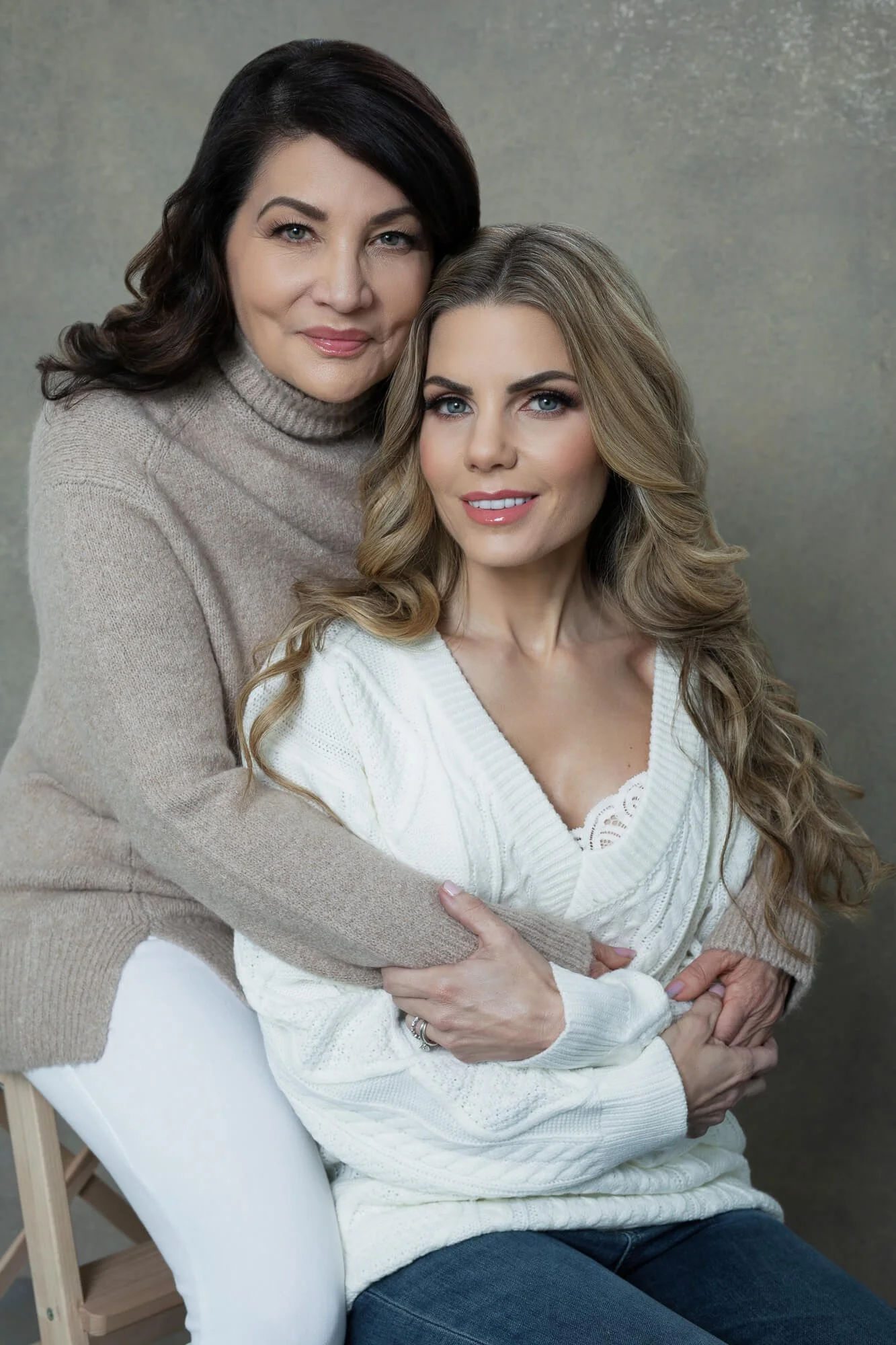 A woman with long, wavy blonde hair sitting on a chair wearing a white sweater, embraced by an older woman with dark hair wearing a beige turtleneck sweater, both smiling.