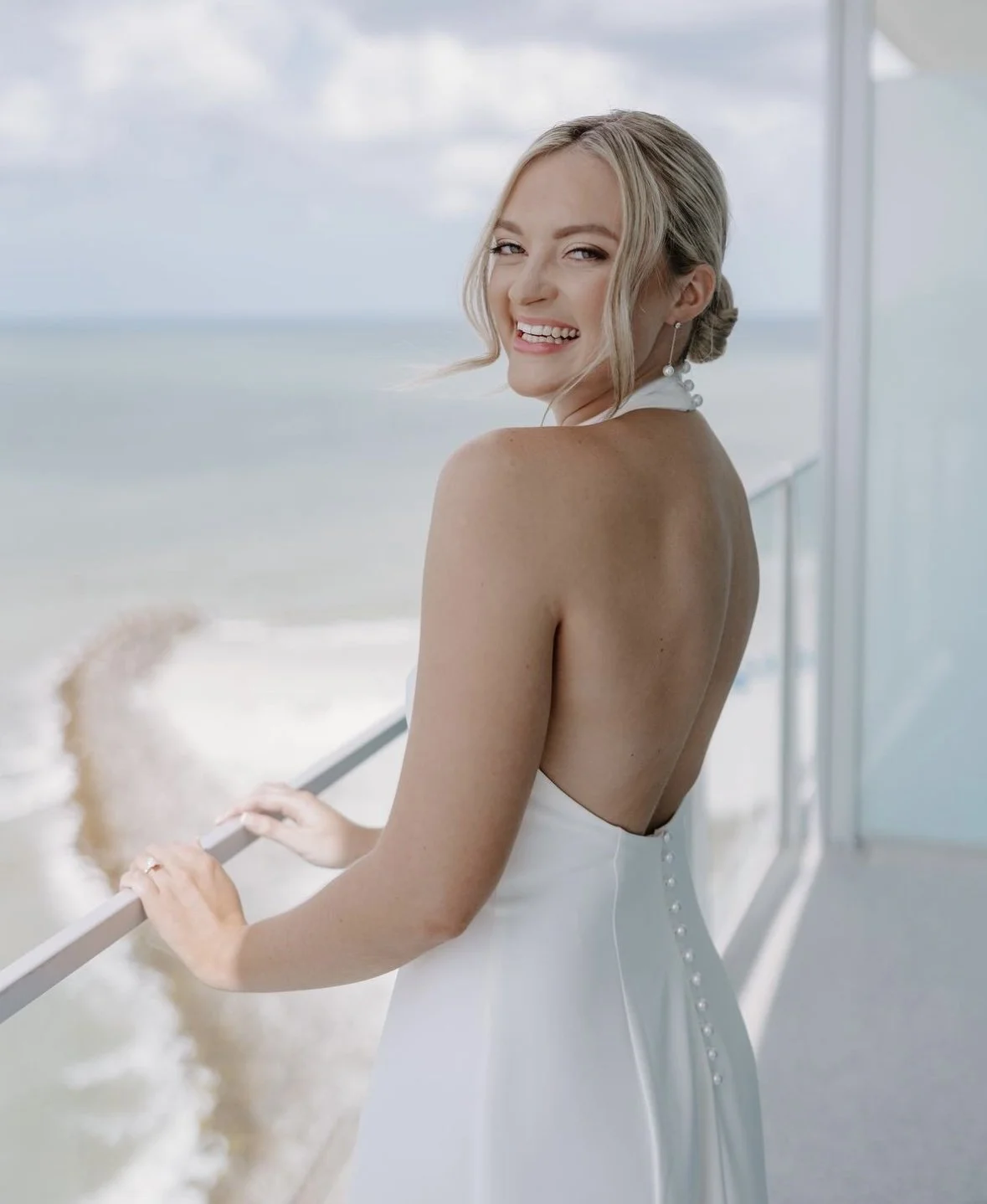 A woman in a white dress with a low back, standing on a balcony near the ocean, smiling and looking over her shoulder.