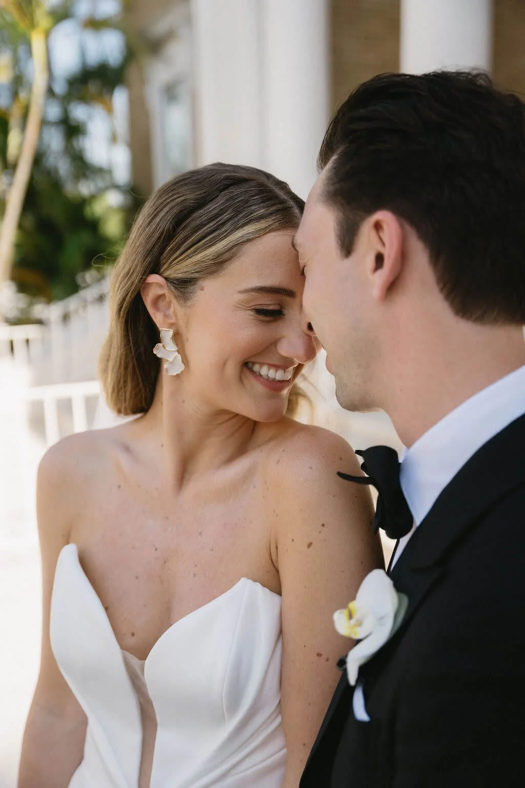 A bride and groom share an intimate moment with their foreheads touching. The bride has shoulder-length brown hair and is wearing a strapless white wedding dress. The groom has dark hair and is in a black tuxedo with a bow tie.