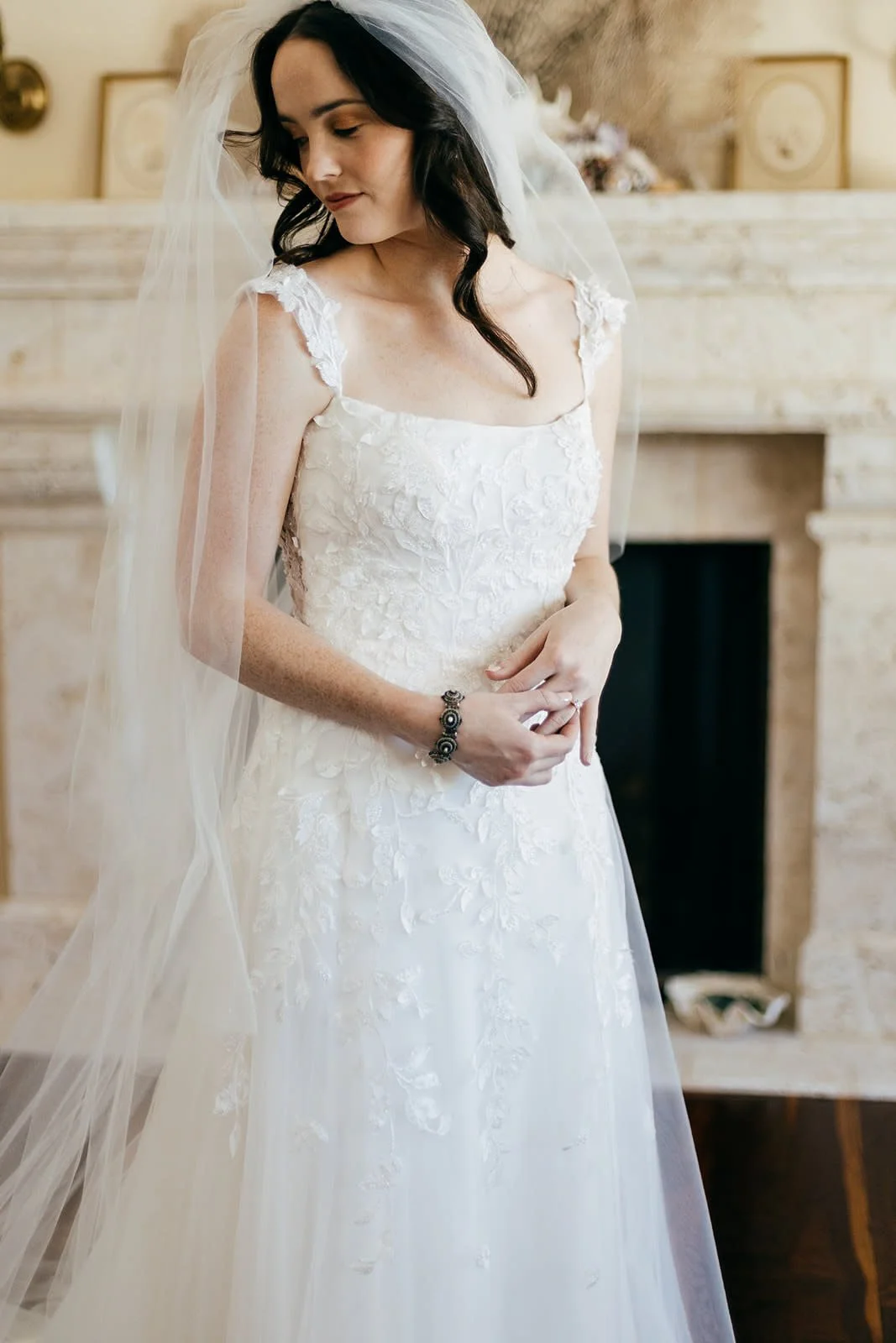 A bride in a white lace wedding gown wearing a veil, standing in a room with a fireplace.